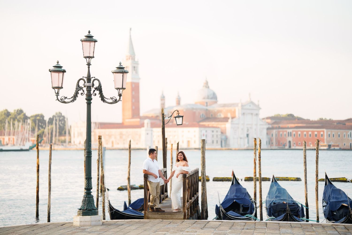 Row of moored gondolas along Venice canal with ornate street lamps and San Giorgio Maggiore church in background.