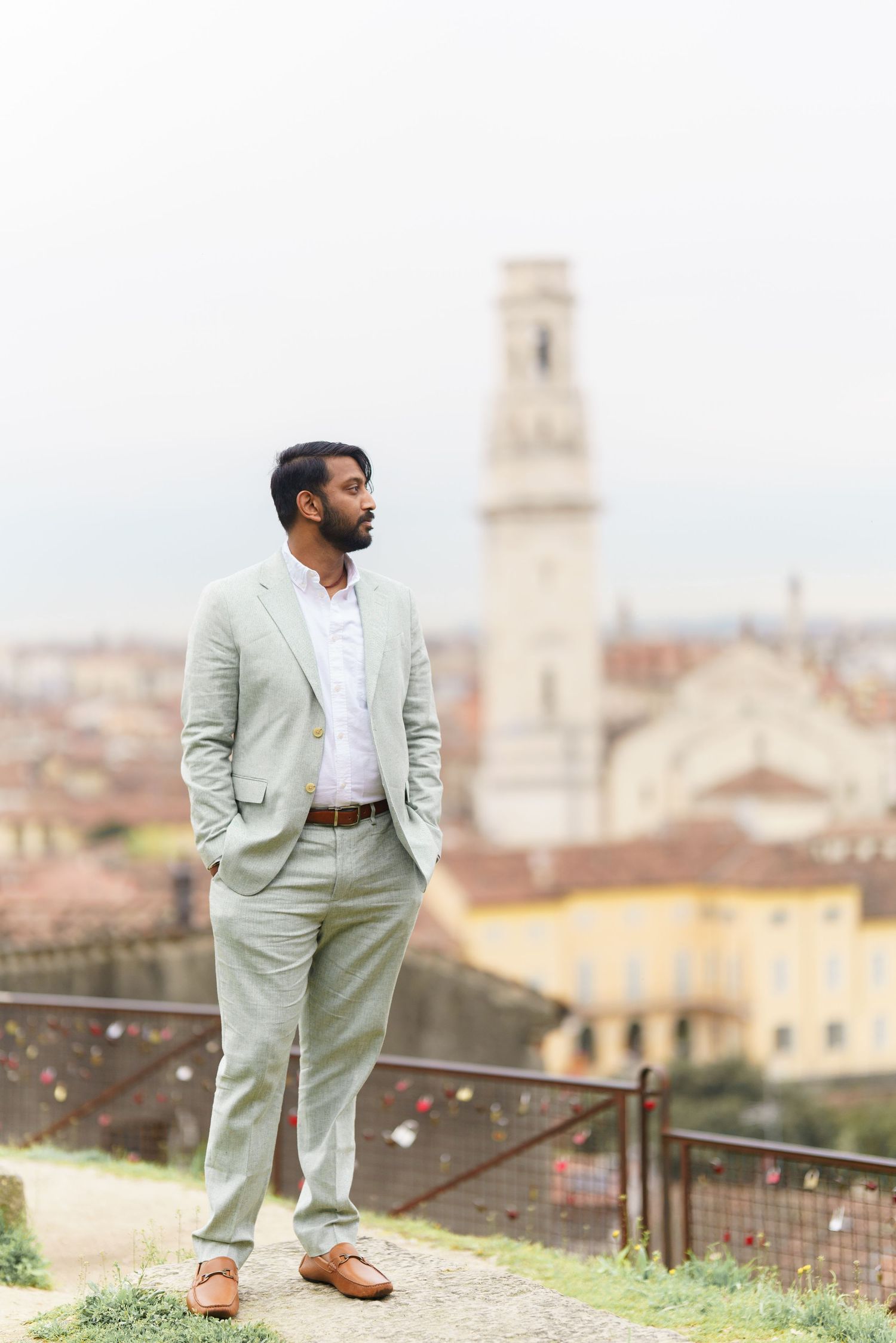 A person in a light mint suit poses against a scenic Italian cityscape backdrop with historic architecture.