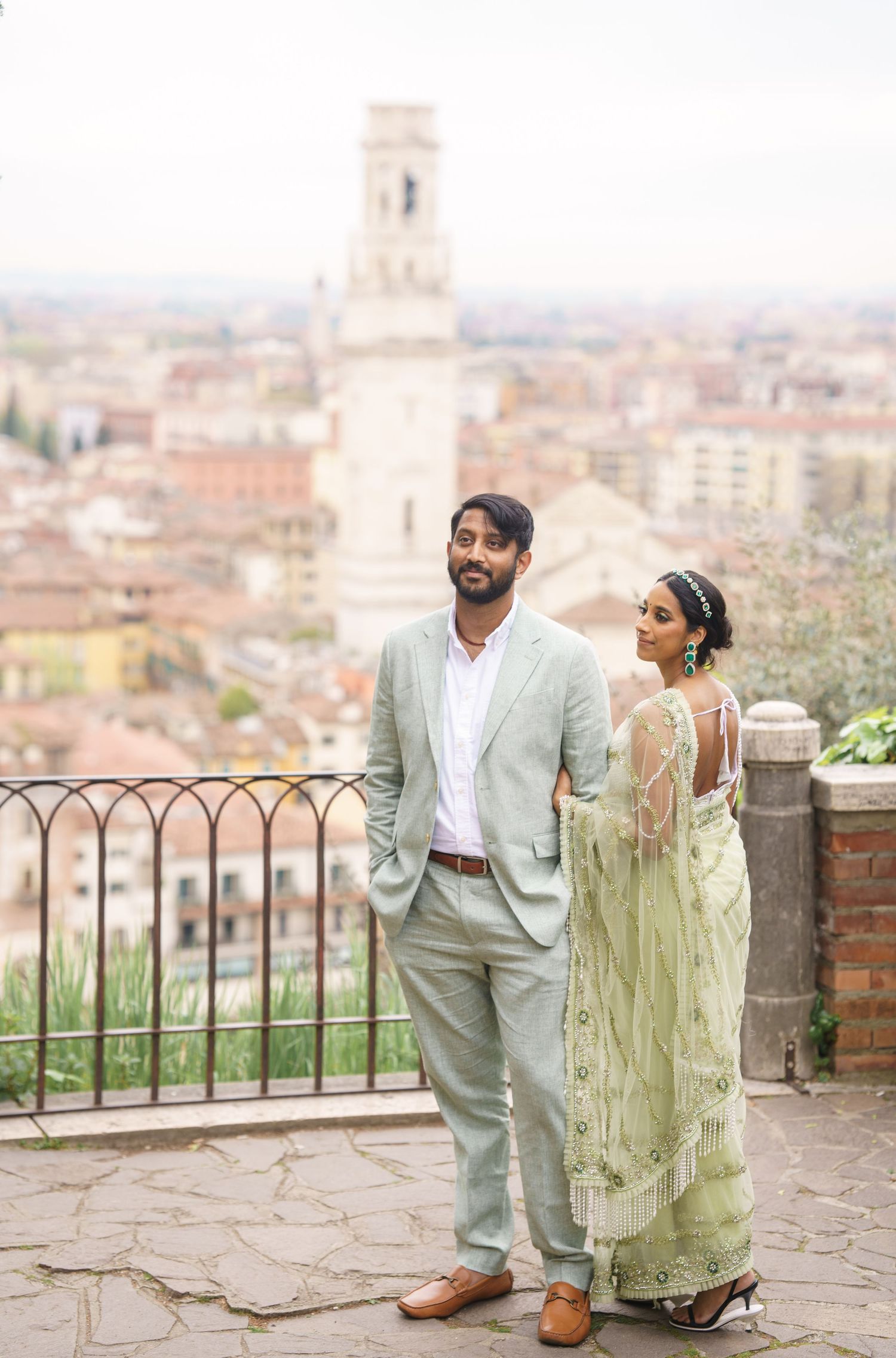 A couple in elegant attire poses against a scenic Italian cityscape backdrop on a stone terrace.