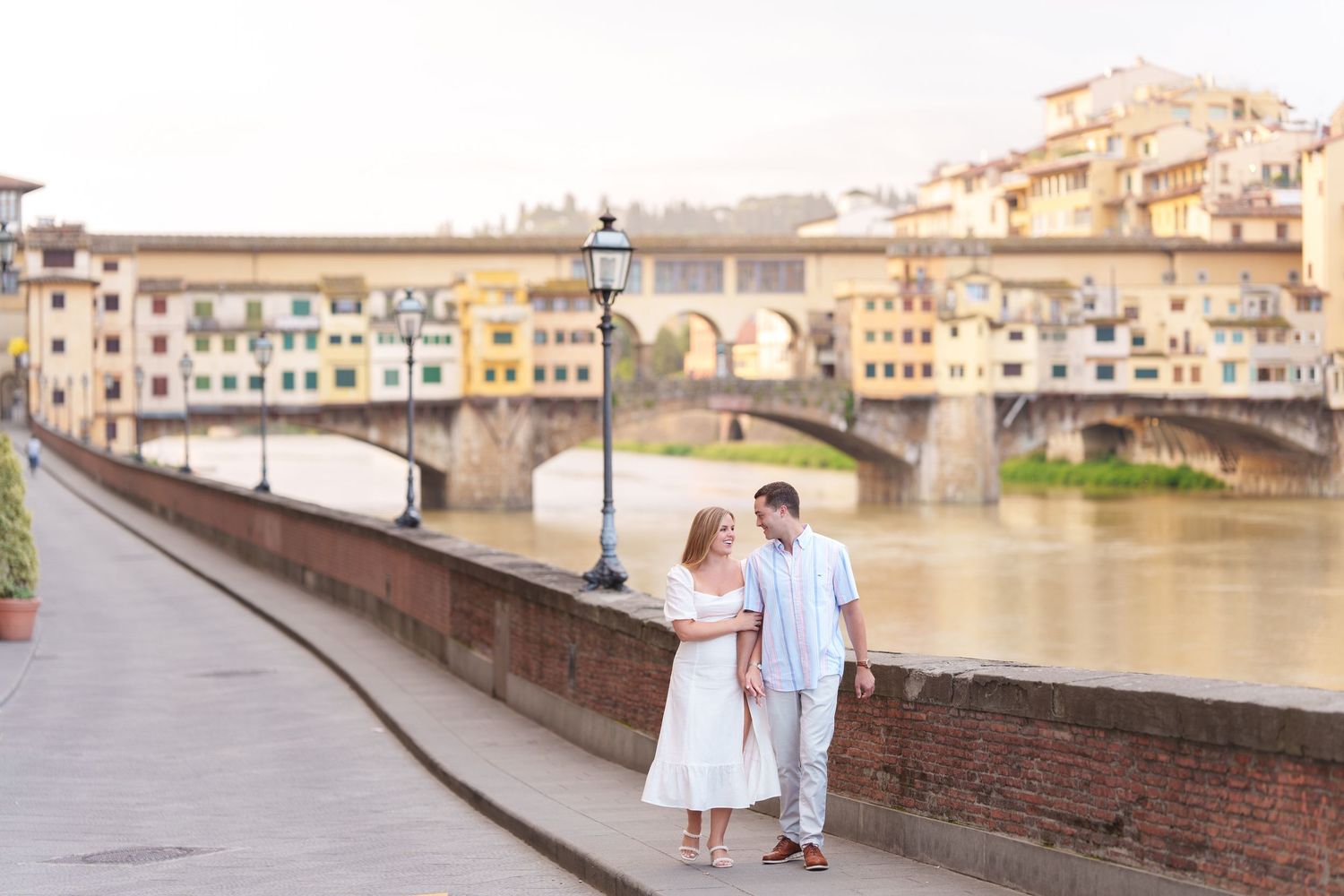 A couple shares an umbrella while strolling along the Arno River with Florence's iconic Ponte Vecchio bridge in the background.