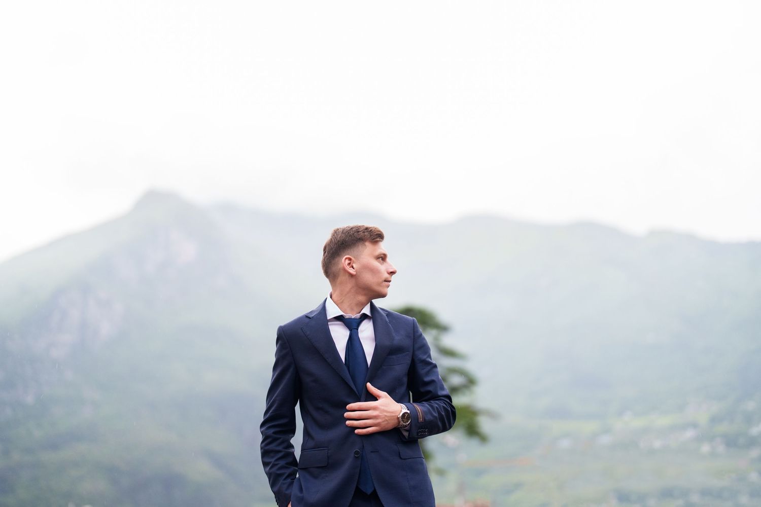 A person in a navy suit stands in front of a misty mountain landscape with a dramatic cloudy sky.