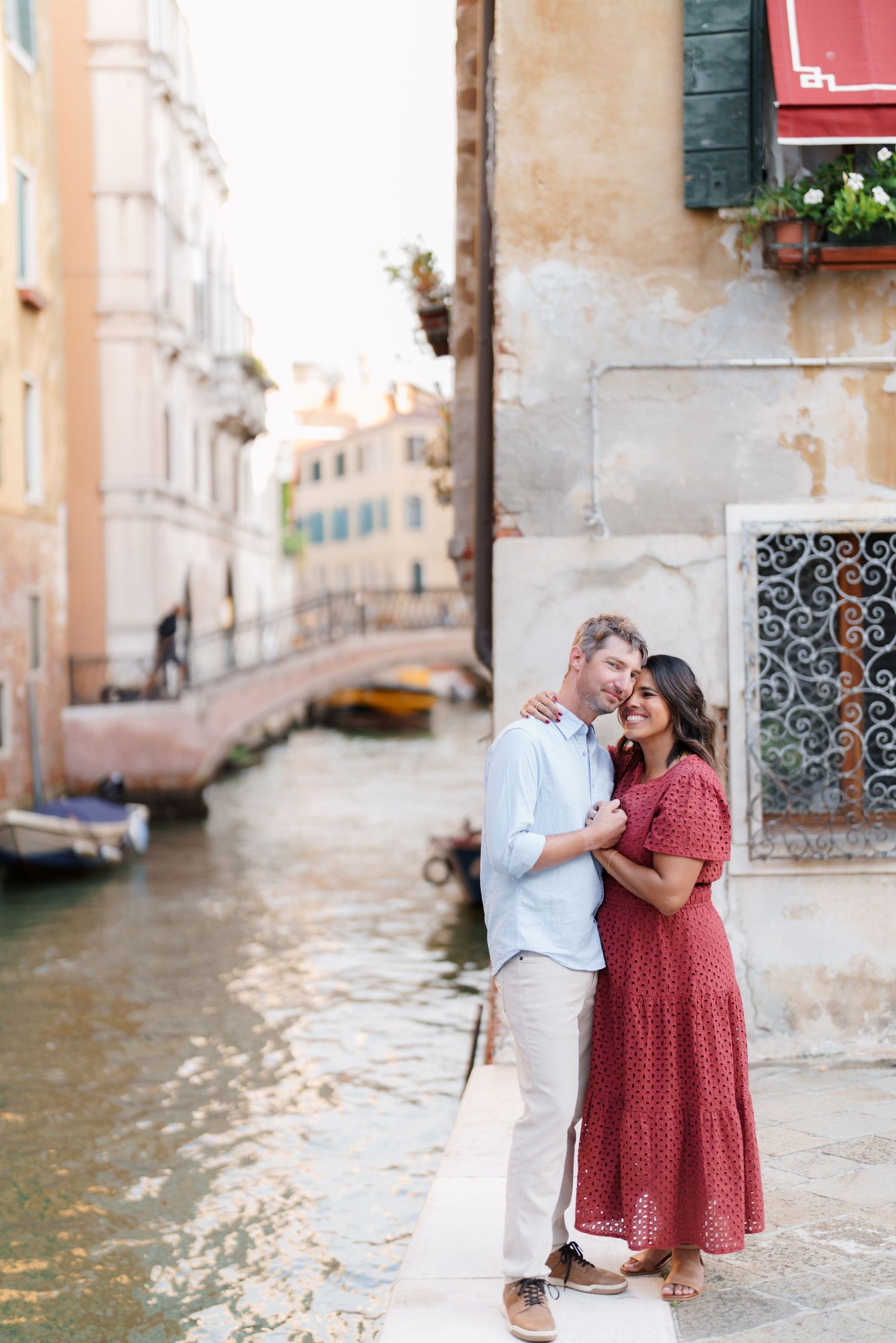 Couple embracing by a Venice canal with colorful buildings and a bridge in the background.