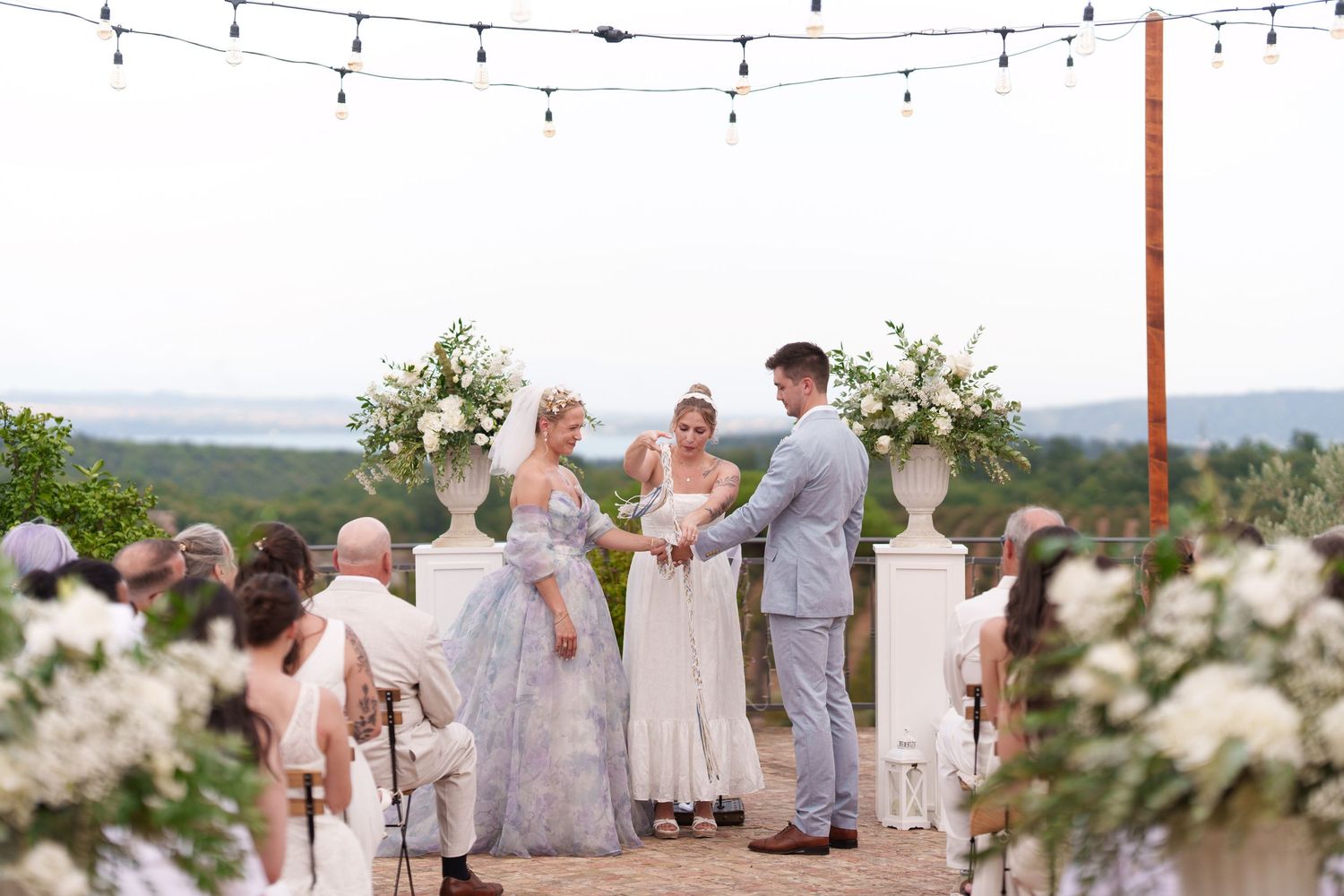 Outdoor wedding ceremony with couple exchanging vows at floral altar, white urn arrangements, string lights, and scenic views.