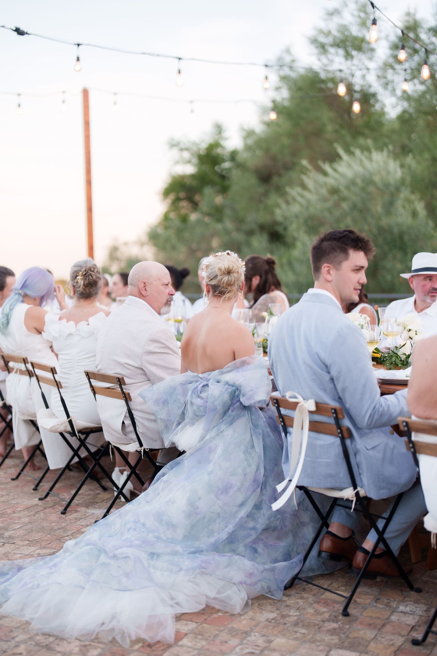 Bride in blue floral gown and groom in light blue suit seated at outdoor wedding reception dinner table.