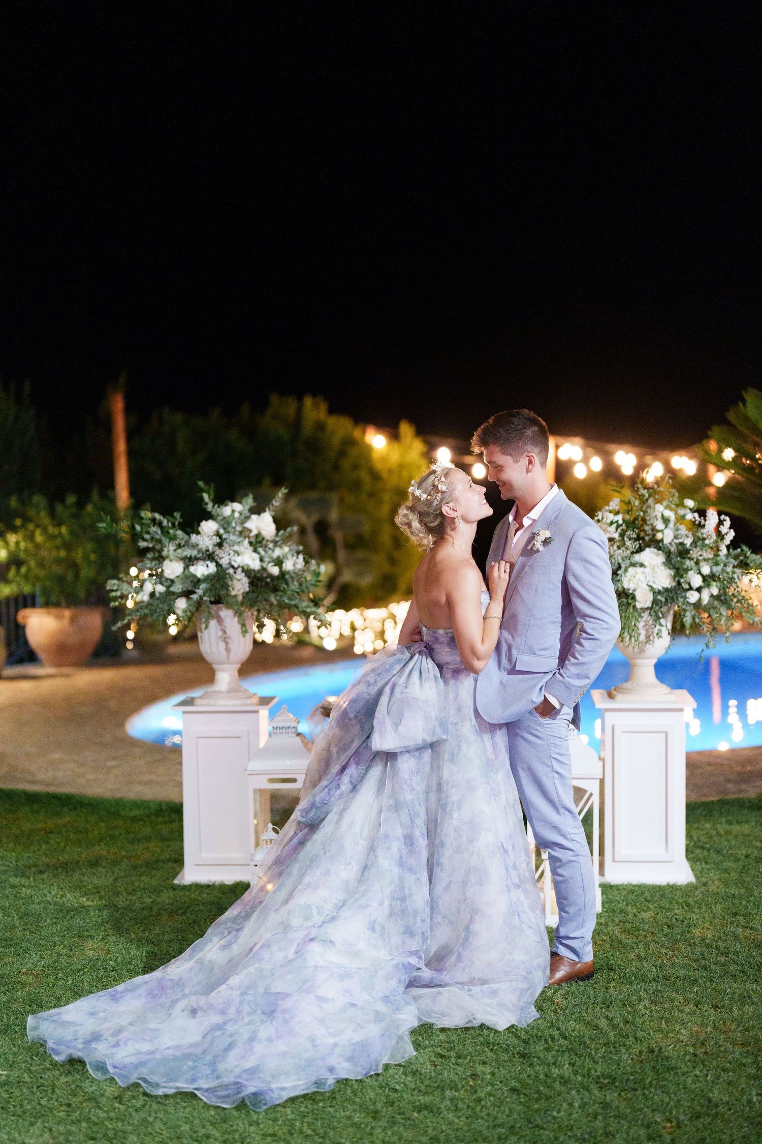 Bride in blue floral gown and groom in gray suit share romantic moment by pool at night wedding.