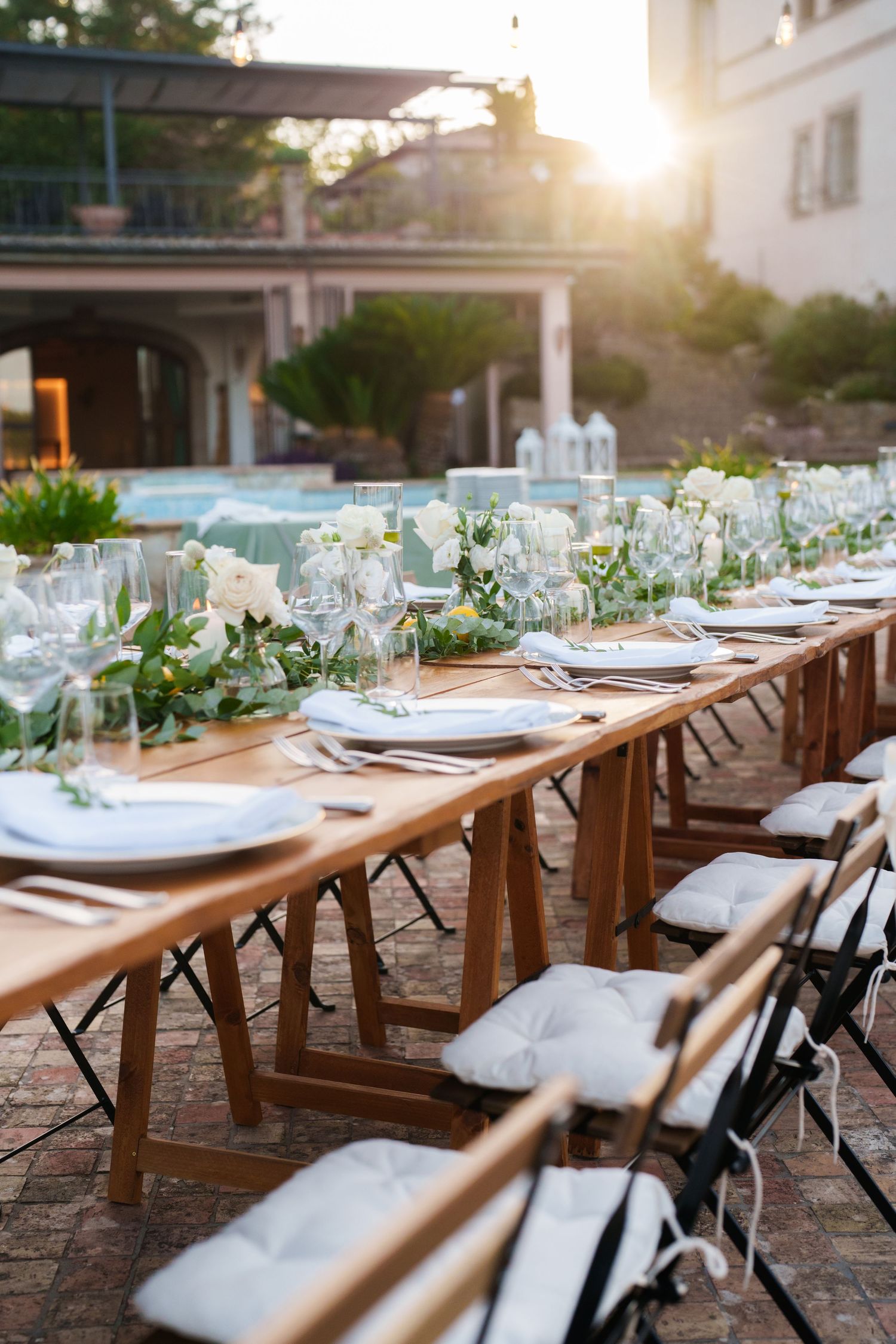Elegant outdoor wedding reception table set with white flowers, glassware, and cushioned chairs in golden sunlight.