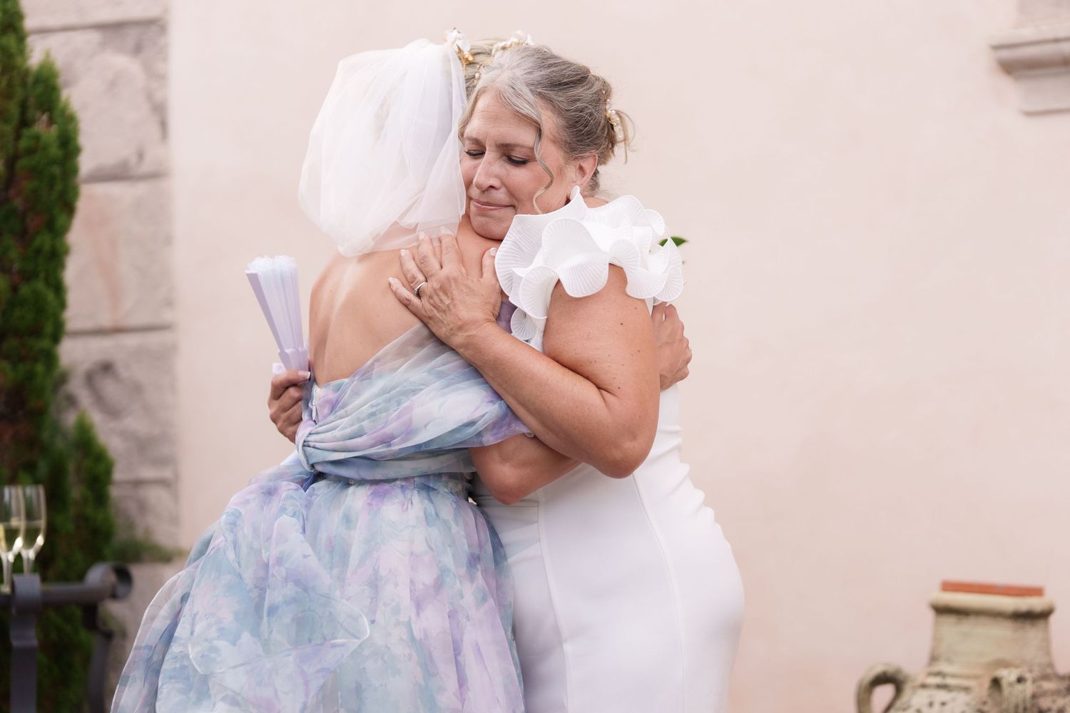 Two women in elegant dresses share a heartfelt hug at a wedding celebration outdoors.