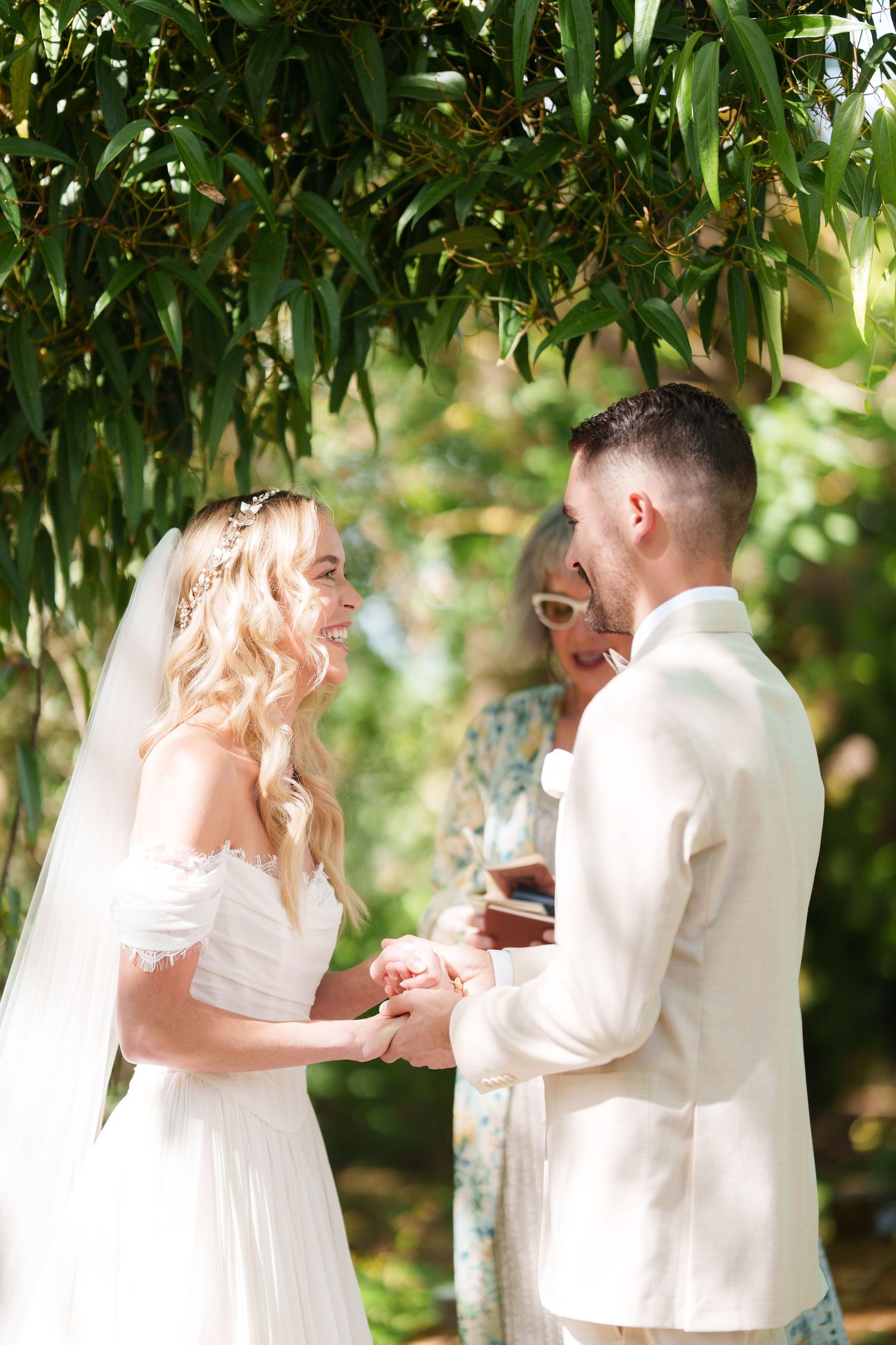 Bride and groom exchange vows outdoors under green trees, officiant in background, sunny garden wedding ceremony.