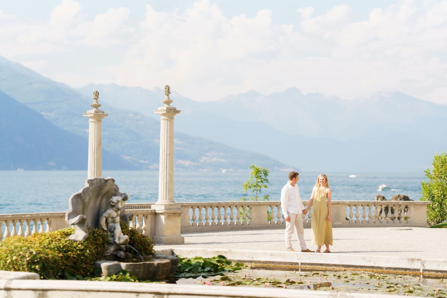 Couple strolling on elegant lakeside terrace with marble columns, fountain sculpture, and mountain backdrop.