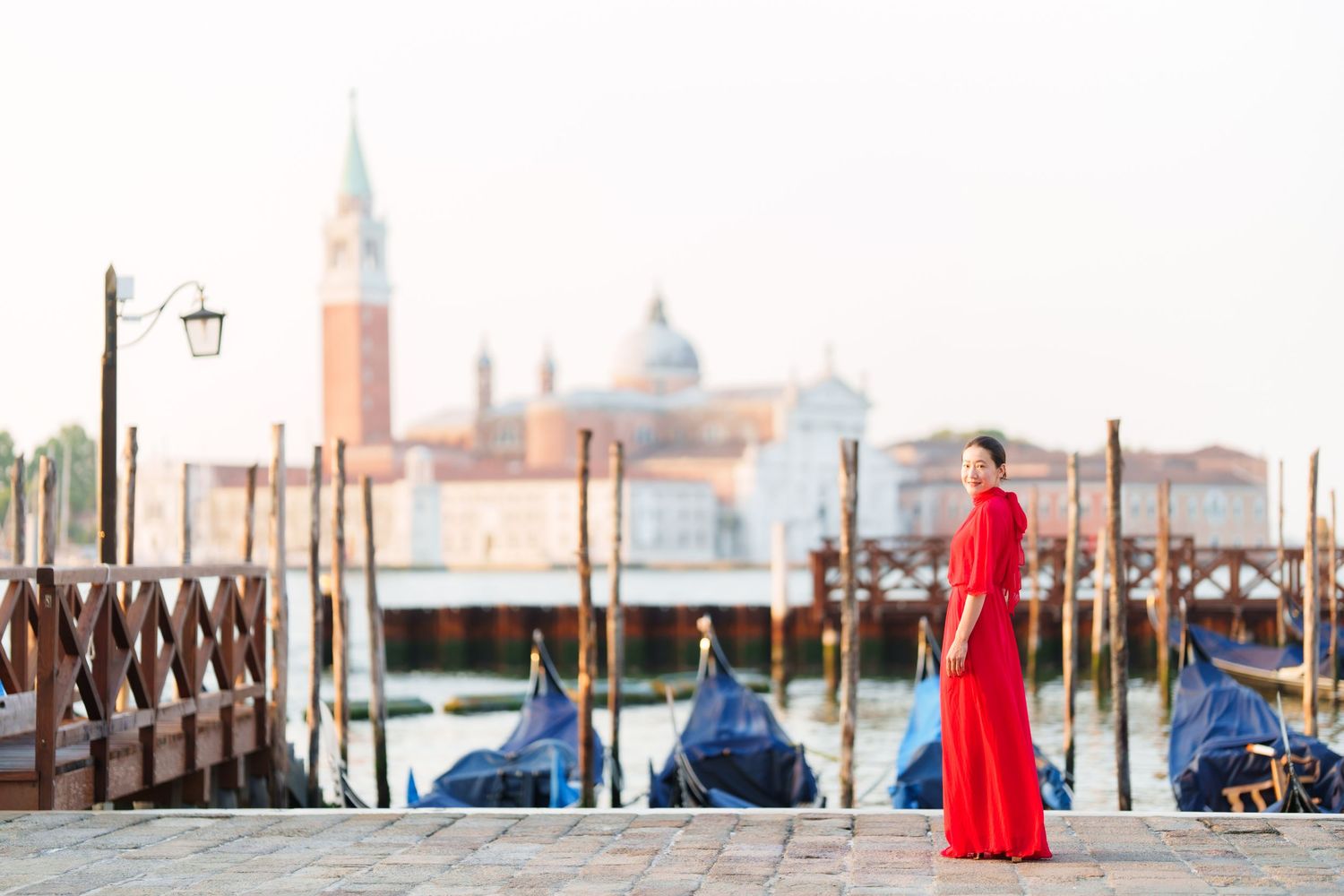 Woman in red dress stands by Venice gondola dock with San Giorgio Maggiore church in background.
