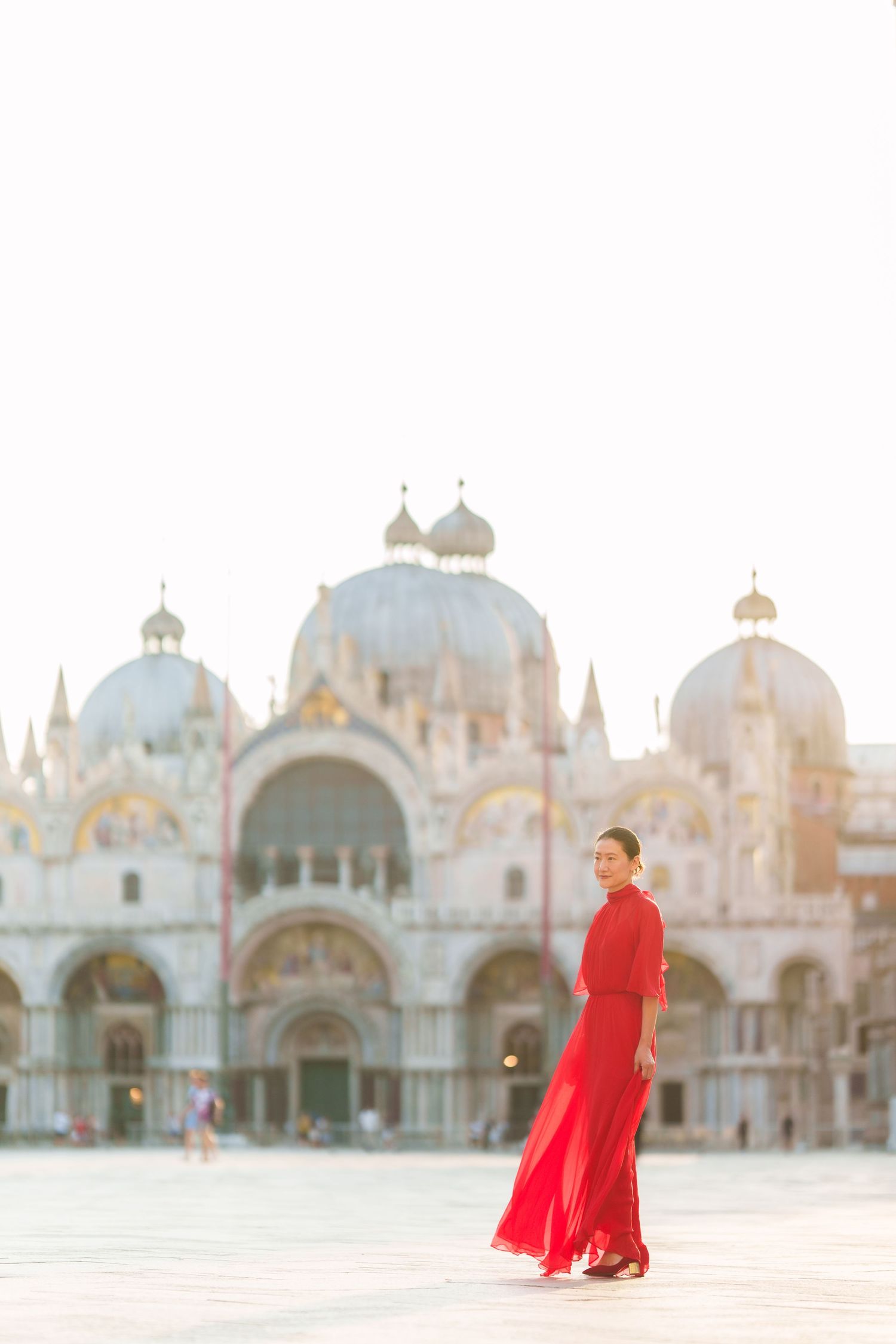 Woman in red dress walking in St. Mark's Square, Venice, with the iconic Basilica di San Marco in the background.