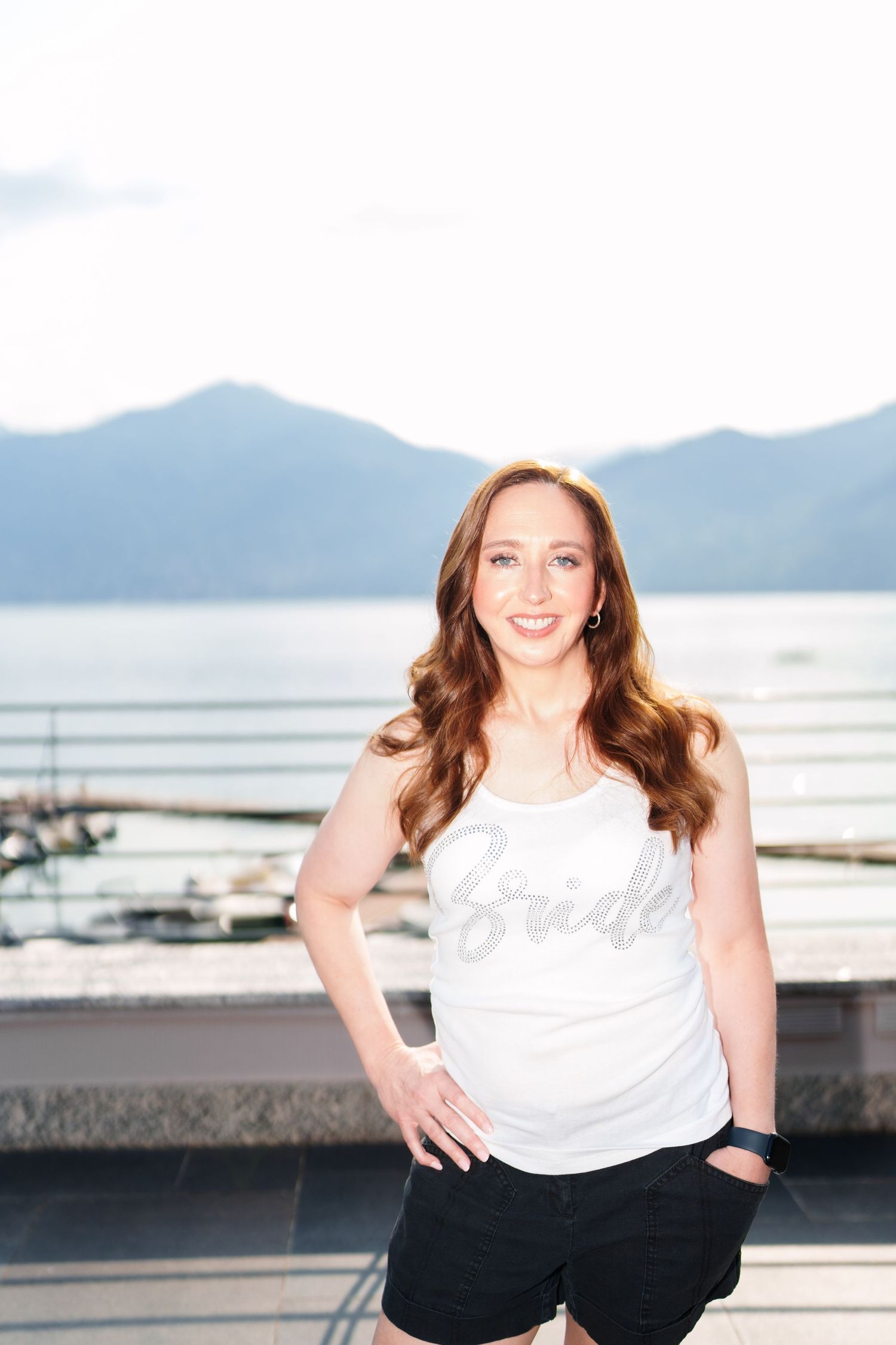 Smiling bride in white tank top and black shorts posing outdoors near a lake with mountain views.