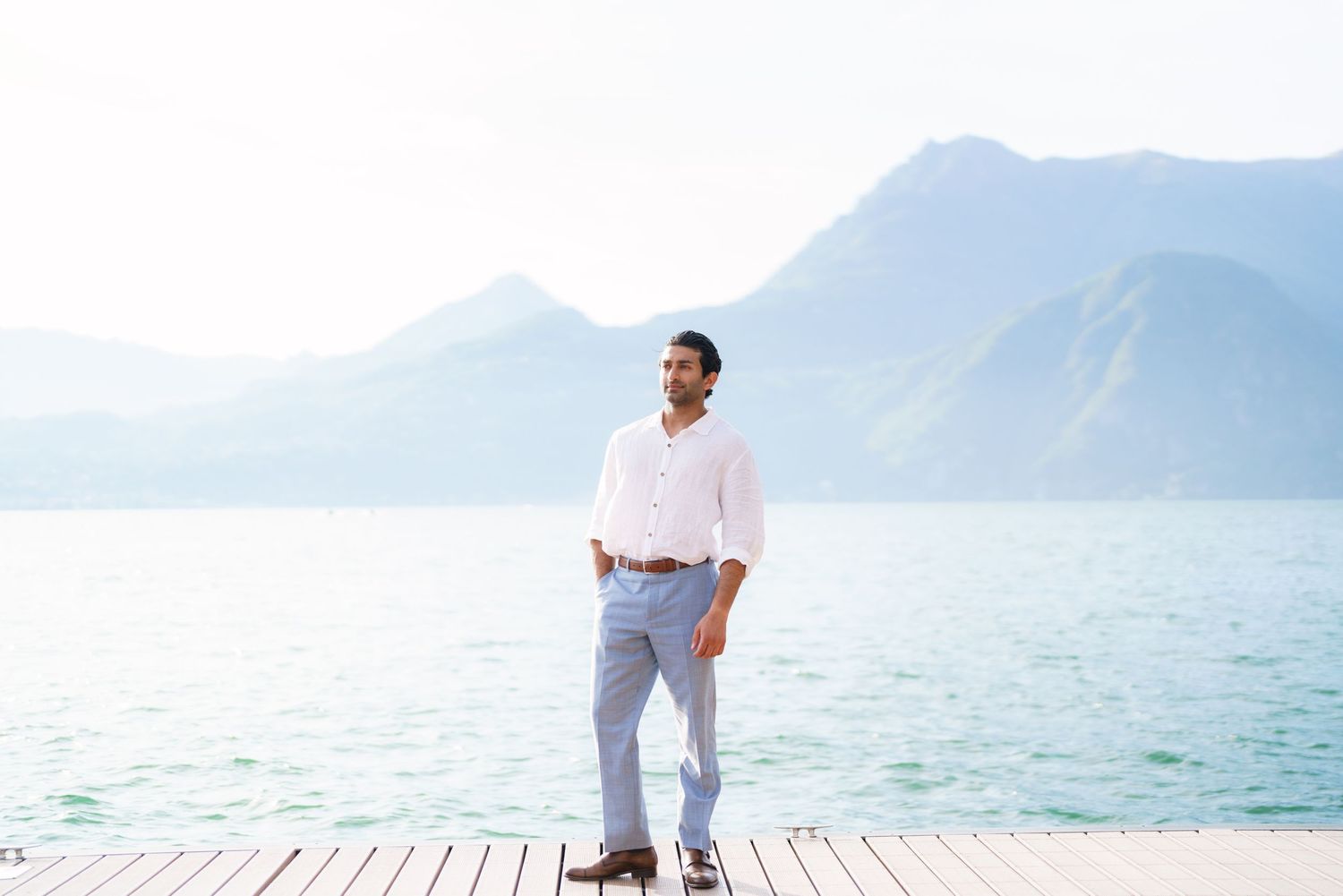 Man in white shirt and gray pants standing on a wooden dock with mountains and turquoise lake in background.