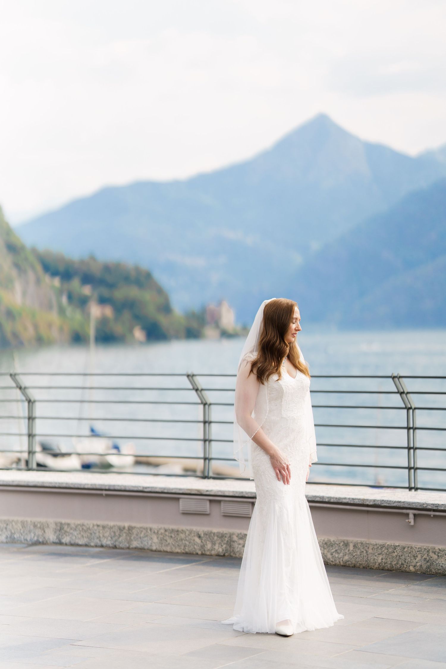 Bride in white mermaid gown stands on lakeside terrace with mountain backdrop on her wedding day.