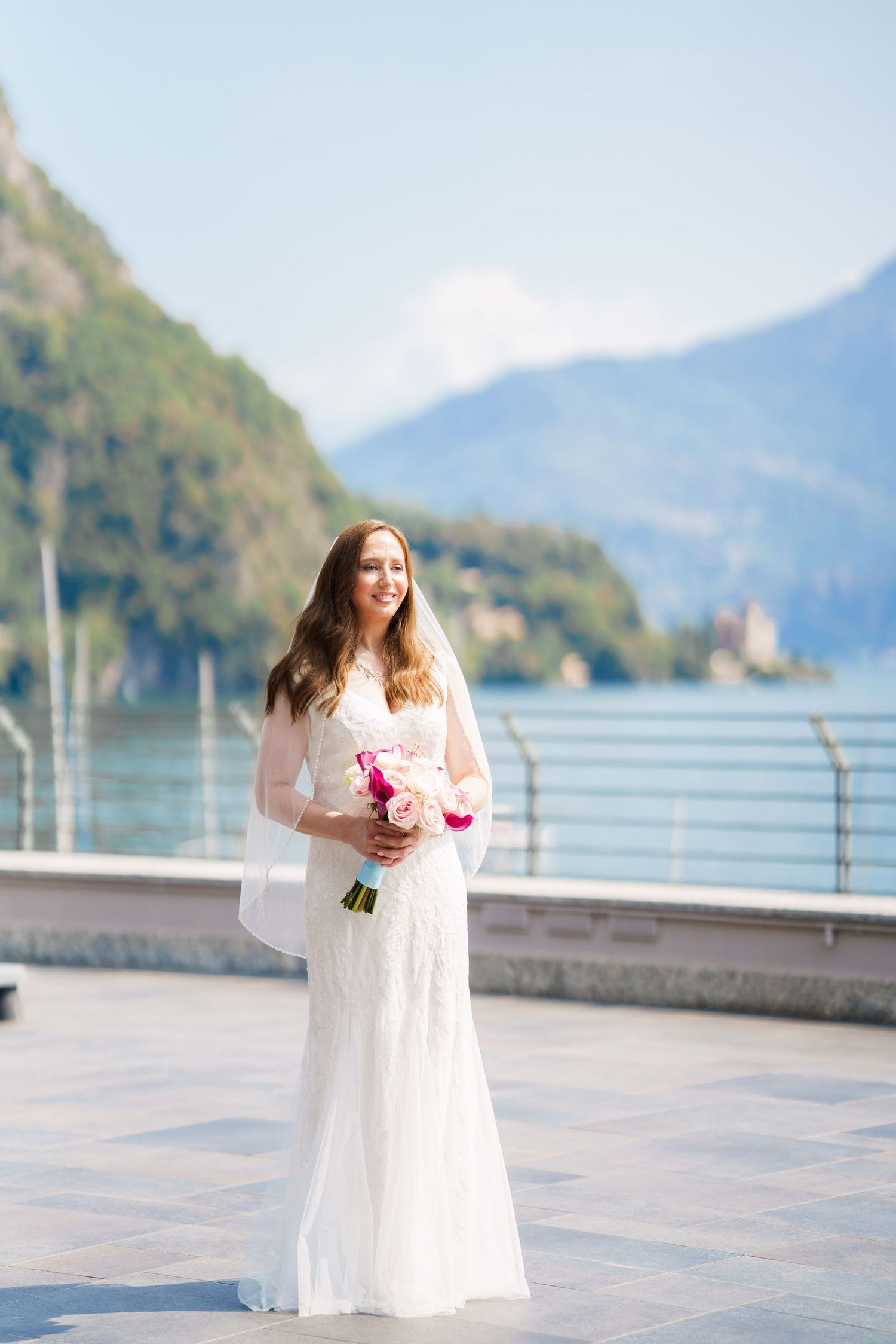 Smiling bride in white lace gown holding pink bouquet stands on lakeside terrace with mountain backdrop.
