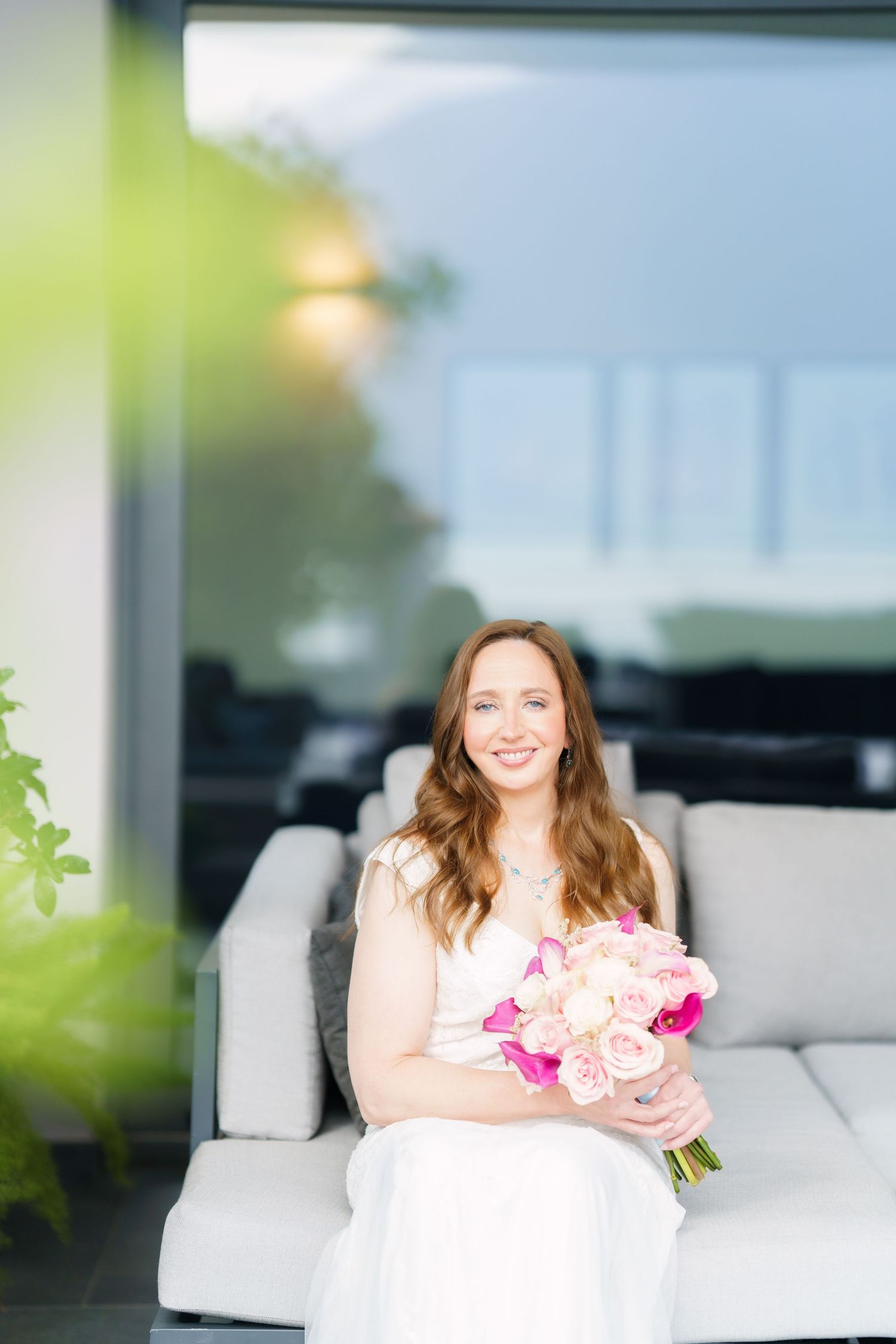Smiling bride in white dress holding pink and blush floral bouquet seated on outdoor sofa.