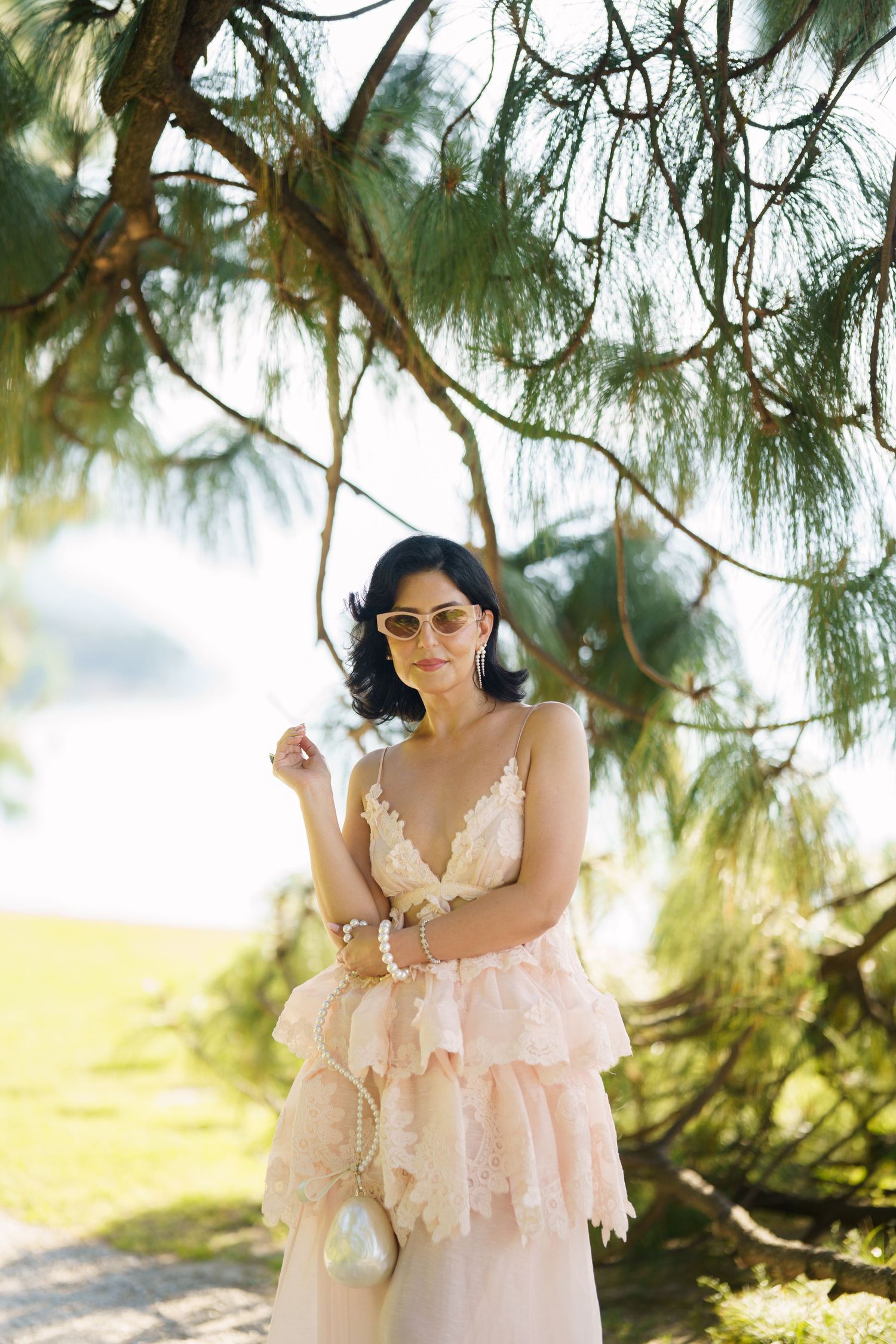 Woman in pink lace dress and sunglasses posing under pine tree branches outdoors.