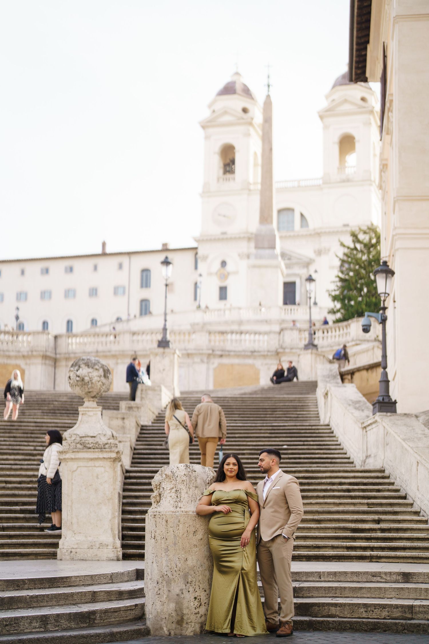 Pregnant couple poses on the Spanish Steps in Rome, woman in green gown, man in beige suit, church visible behind.