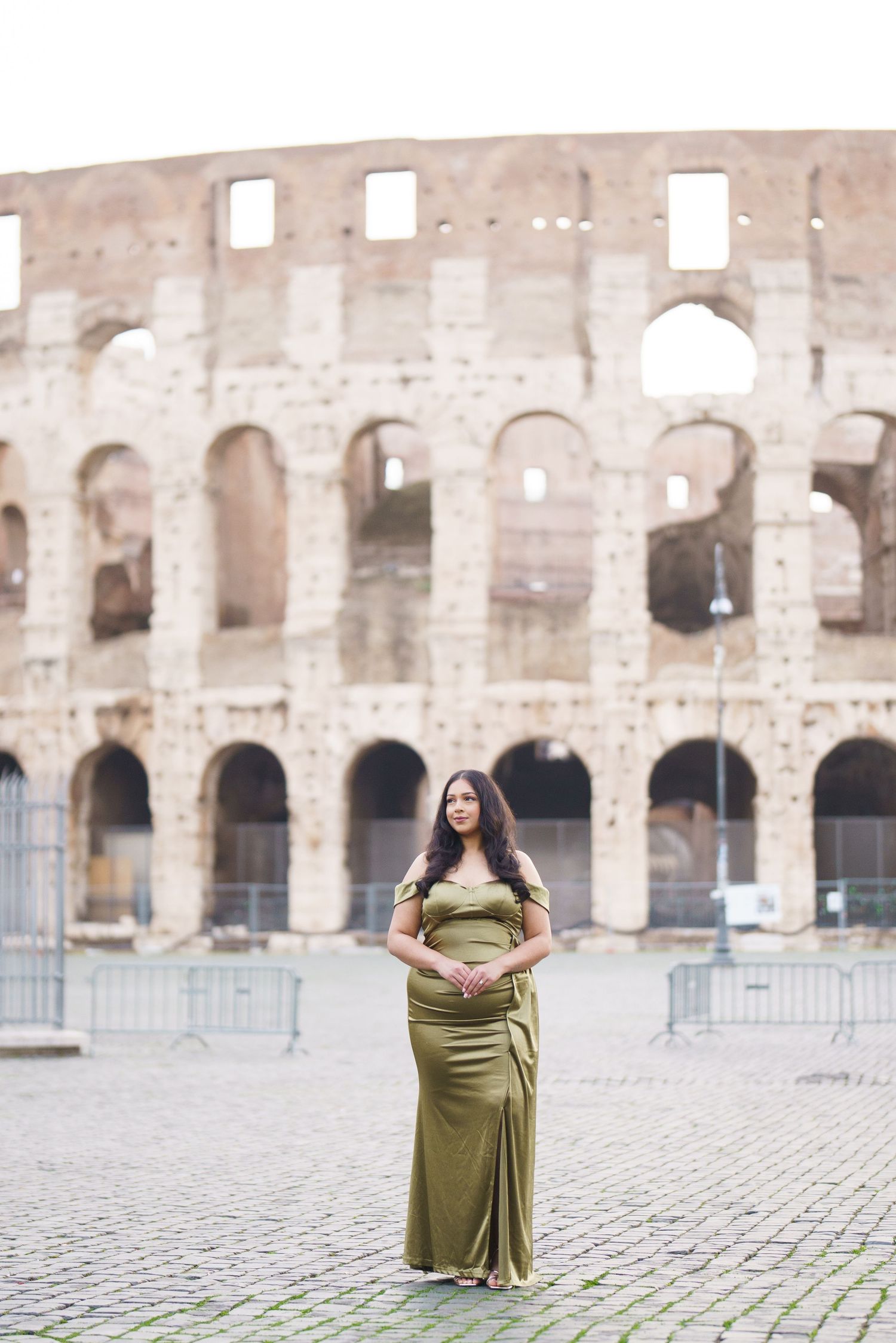 Woman in olive green gown posing in front of the Roman Colosseum on cobblestone plaza.