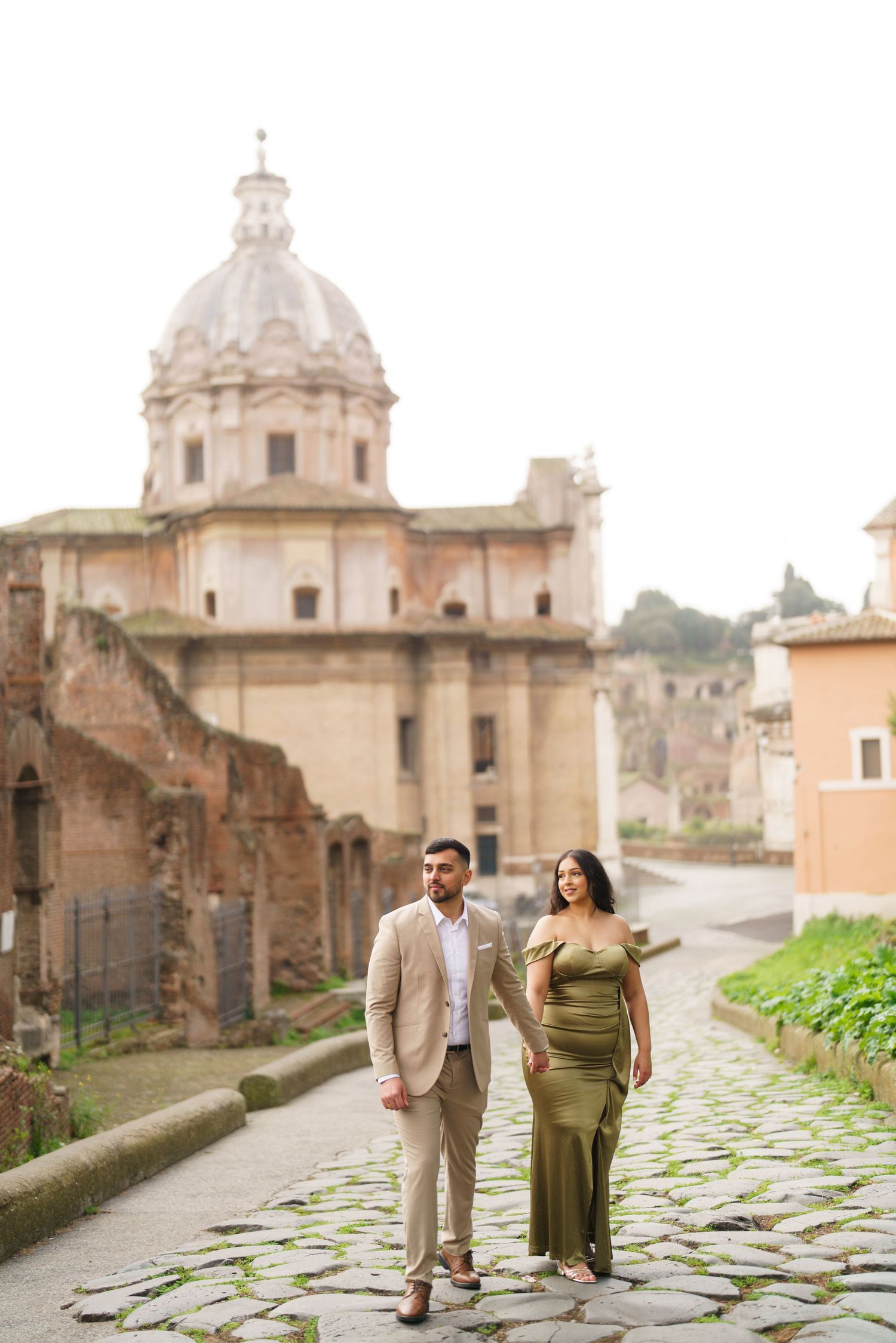 Couple in elegant attire walking on cobblestone path with ancient Roman ruins and domed church in background.