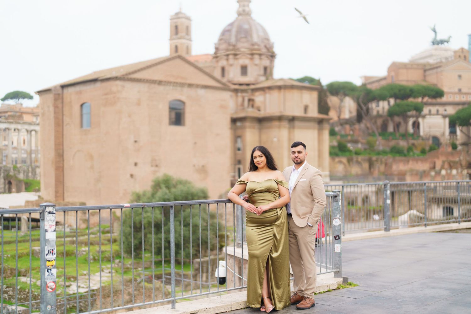Couple posing in elegant attire before ancient Roman ruins and domed church in Rome, Italy.