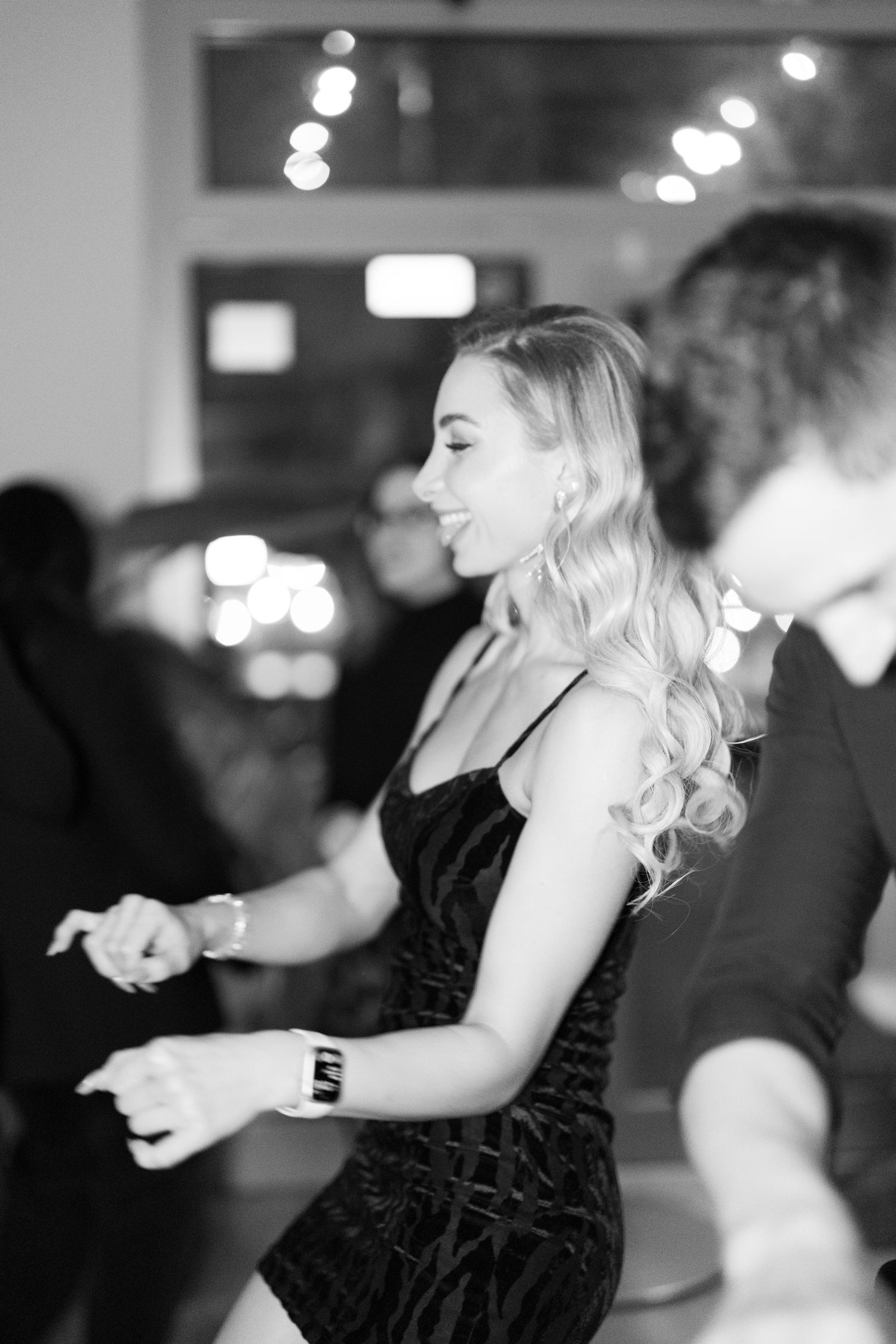 Black and white photo of a smiling blonde woman dancing in a black dress at an elegant event.