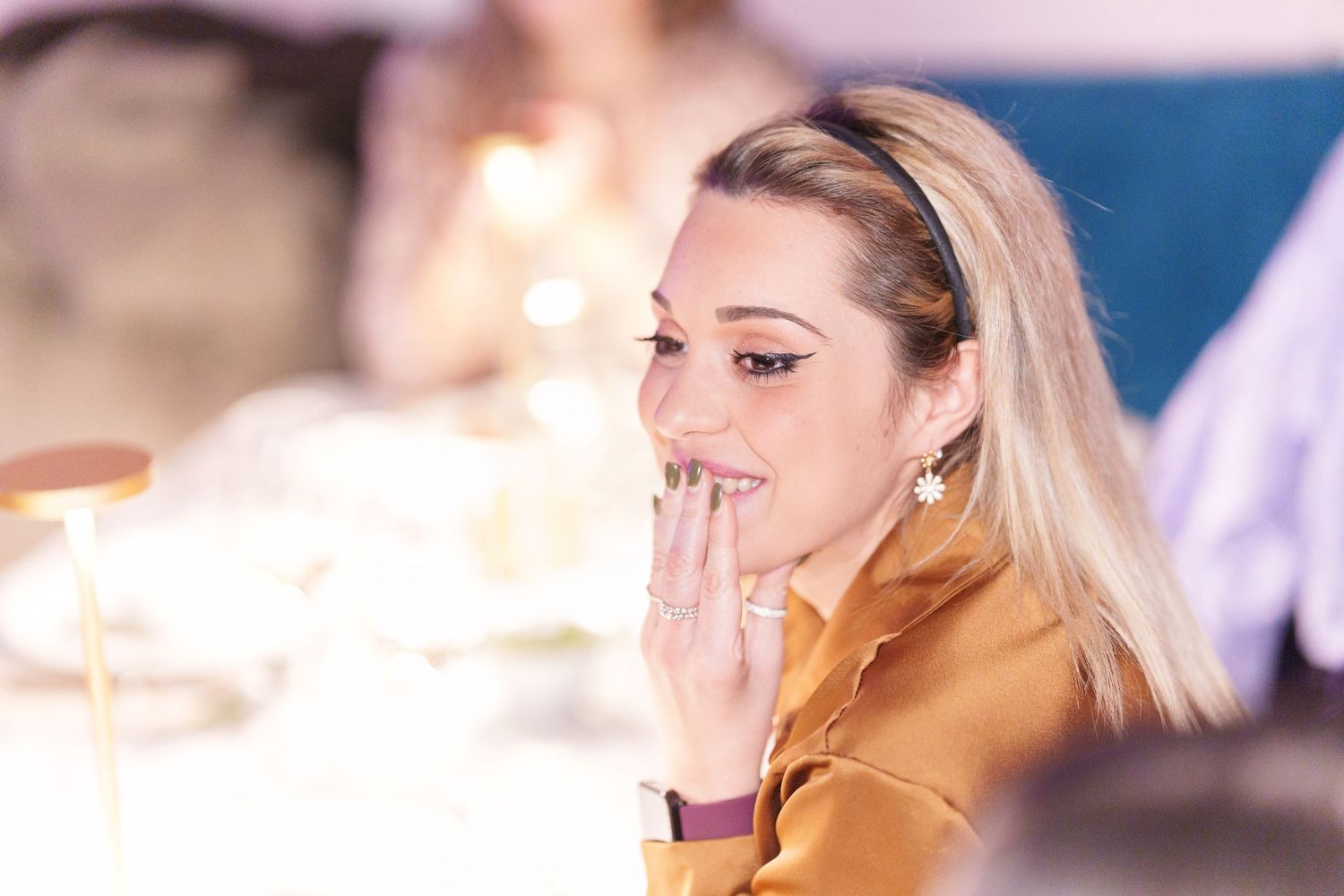Blonde woman in golden top smiling at a dinner event, touching her lips elegantly.