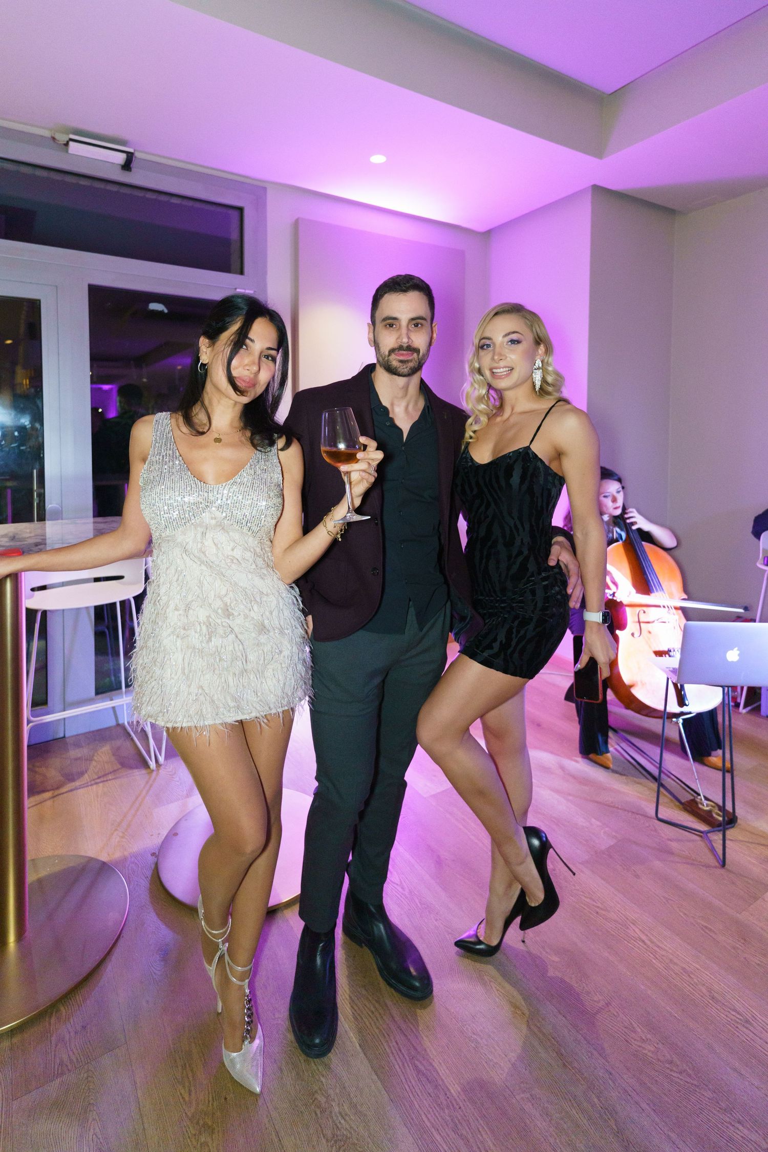 Three elegantly dressed party guests pose together at an upscale event, with purple lighting and a cellist performing in the background.
