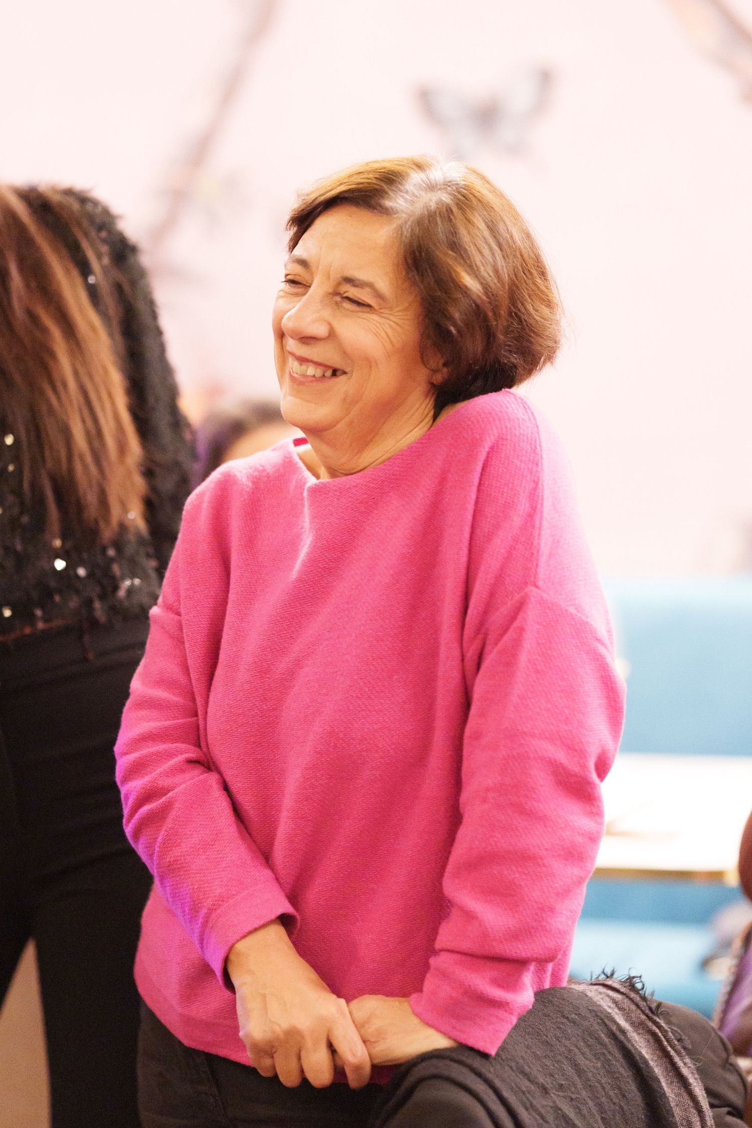Smiling older woman in a bright pink sweatshirt sitting relaxed in a casual indoor setting.