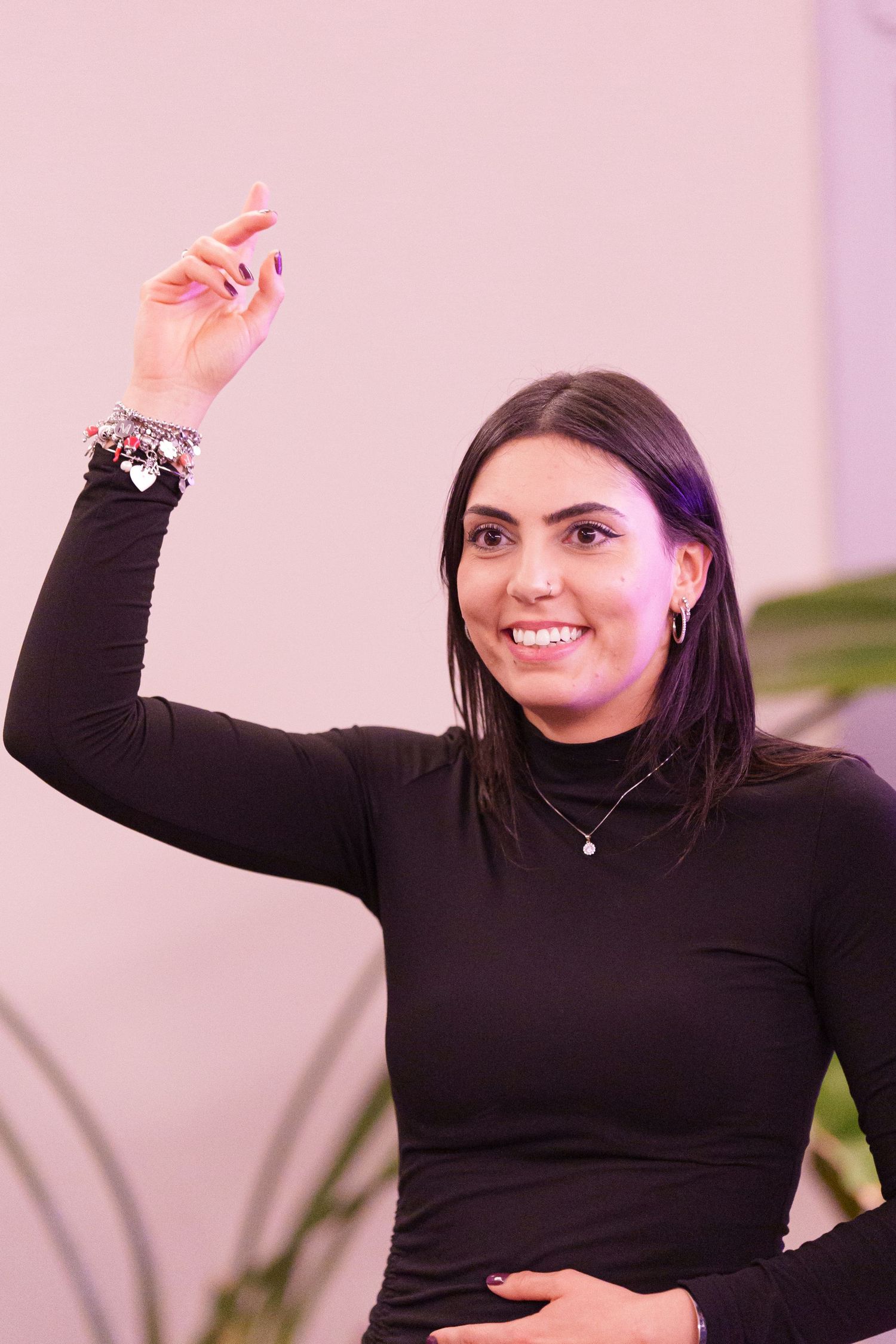 Smiling young woman in black turtleneck raising her hand, wearing charm bracelets and necklace against pink background.