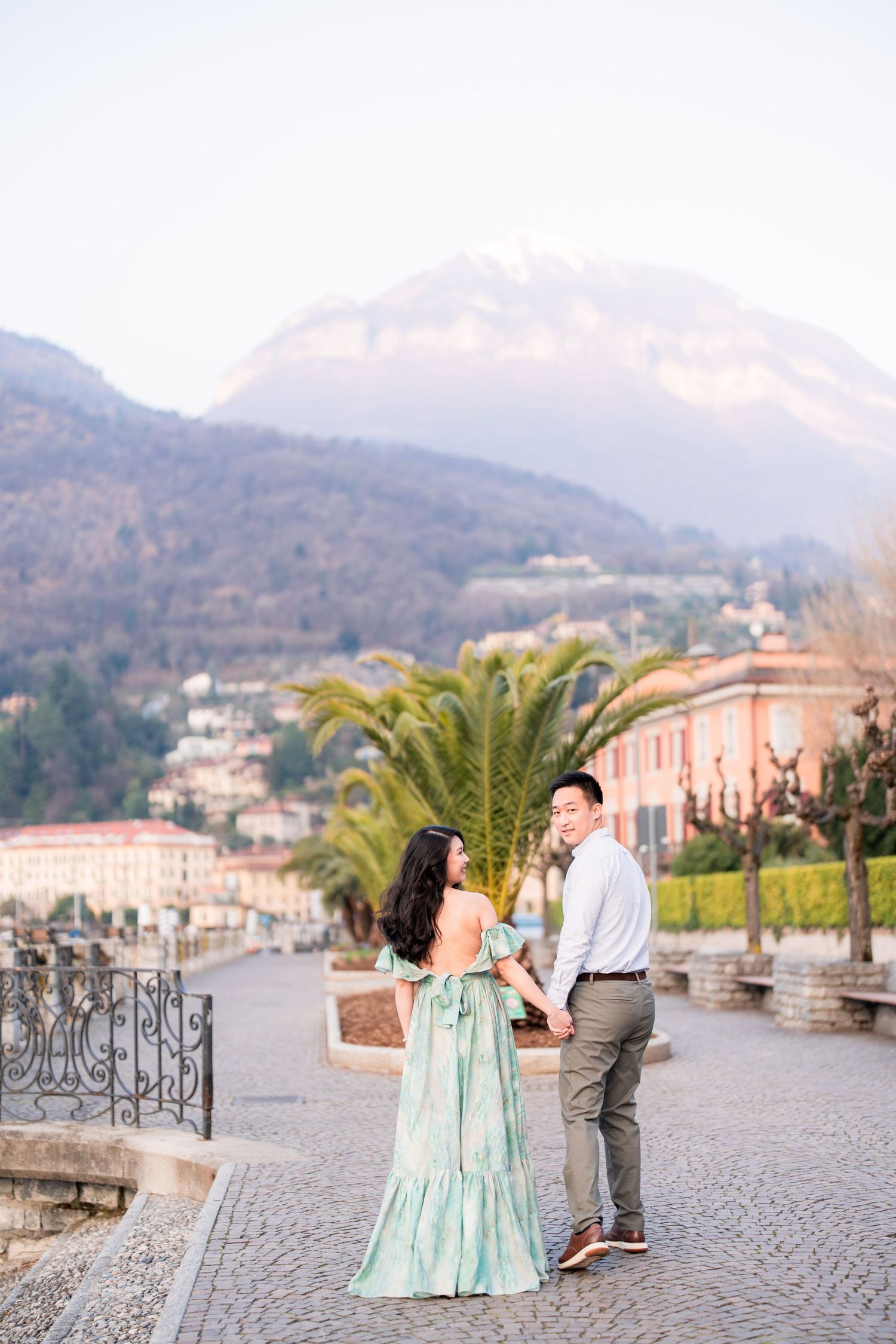 Couple holding hands on a cobblestone promenade with mountain backdrop, woman in mint green dress, Lake Como Italy.