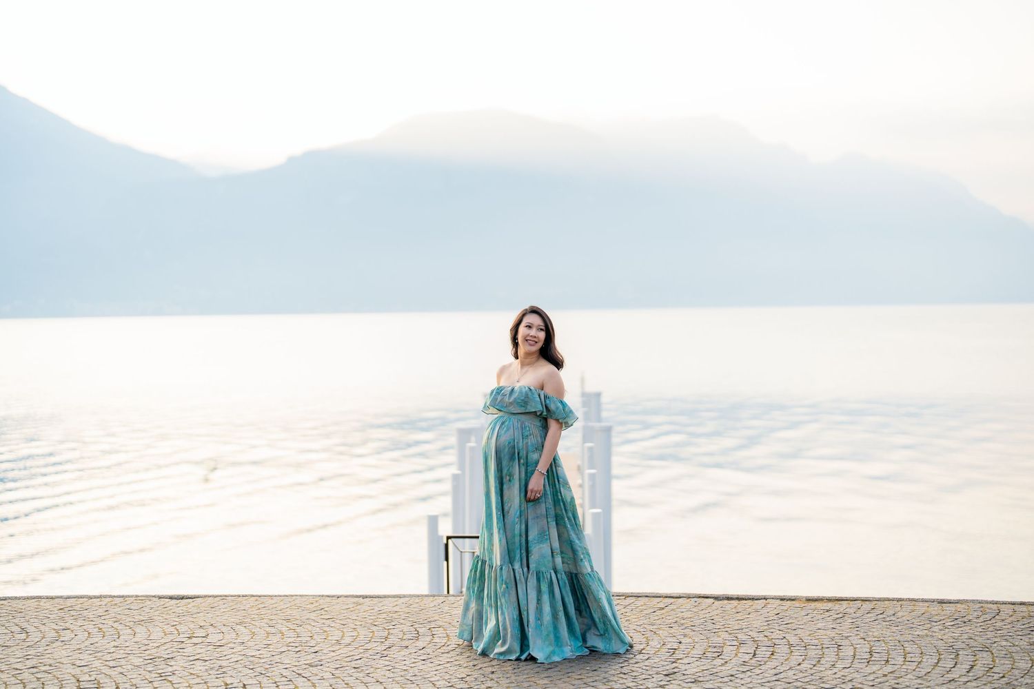 Pregnant woman in teal off-shoulder gown poses by a serene lake with misty mountains in the background.