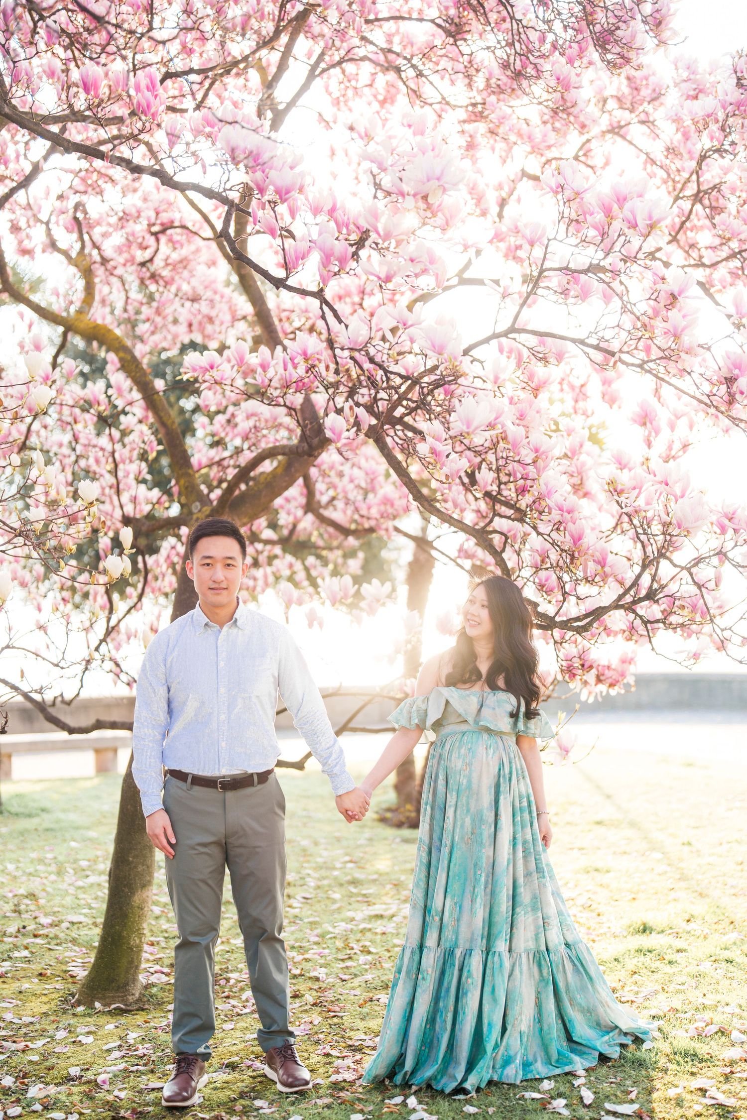 Couple holding hands under blooming pink magnolia tree, woman in teal floral gown, man in blue shirt, spring outdoor portrait.
