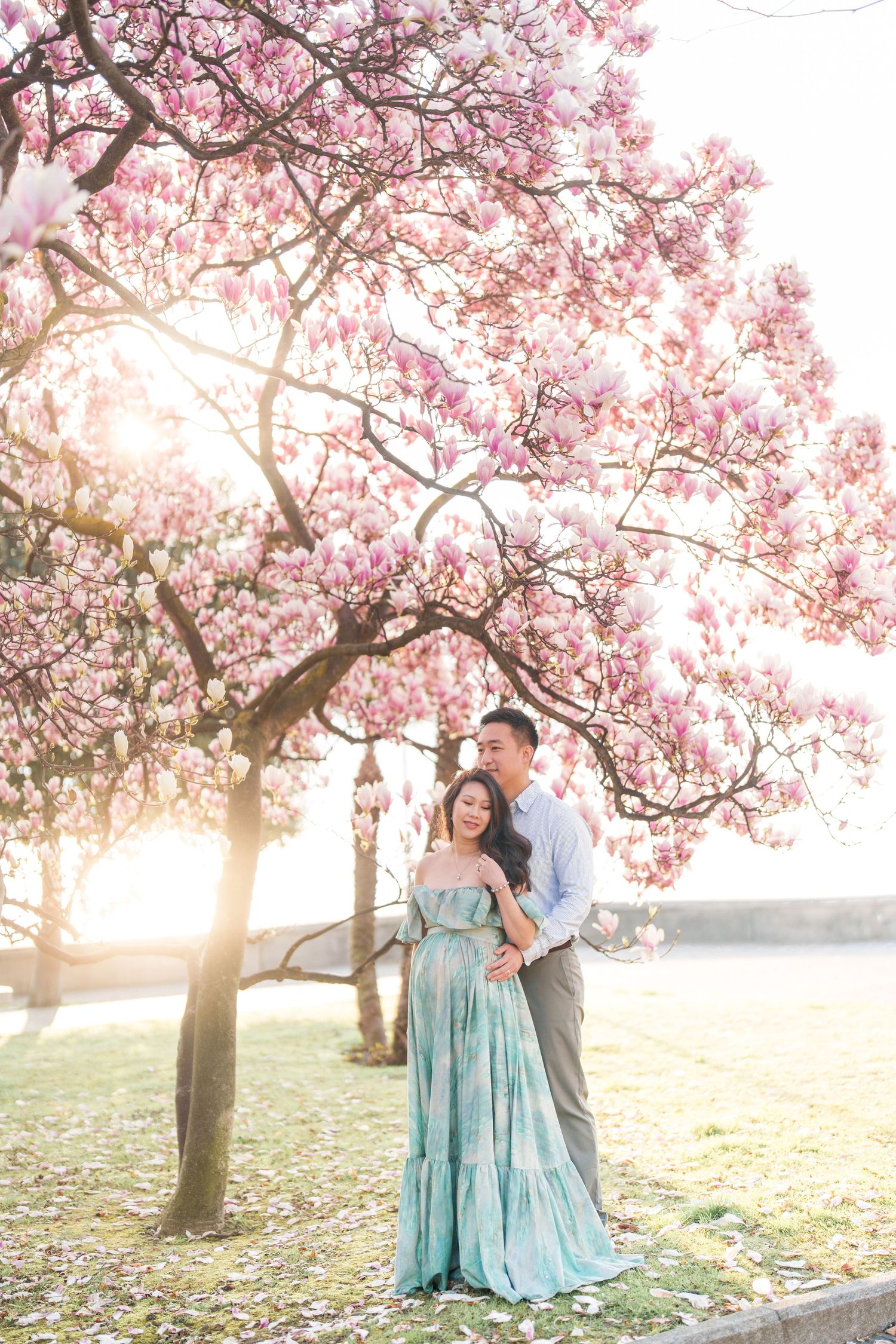 Couple embracing under blooming pink magnolia tree at sunset, woman in flowing teal dress holding baby bump.