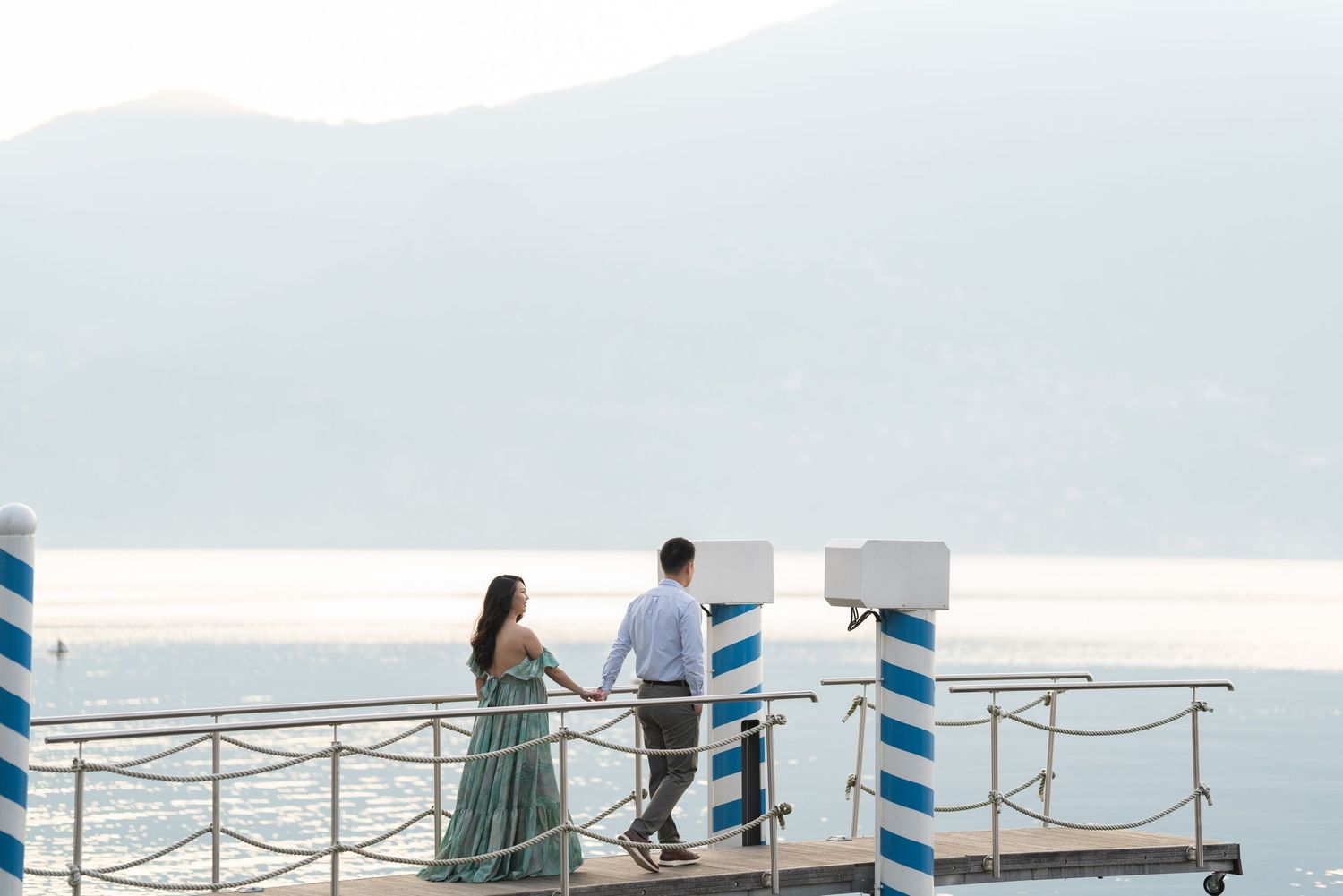 A couple holds hands on a scenic waterfront pier with blue striped posts, mountains and calm sea in the background.