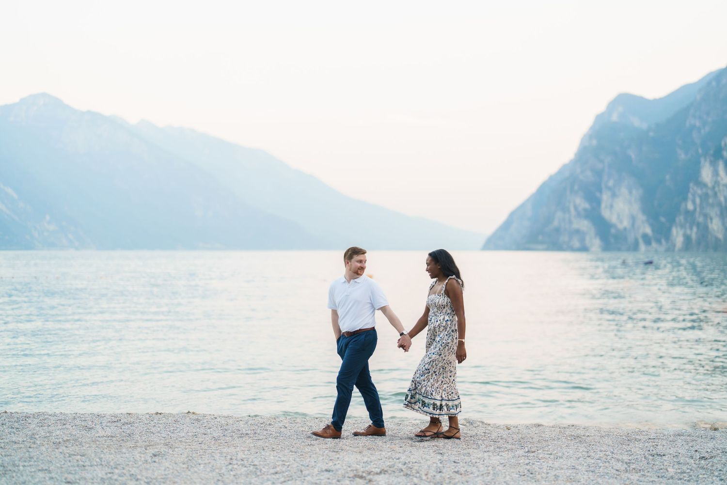 Couple holding hands walking along Lake Garda pebble beach with mountain backdrop.