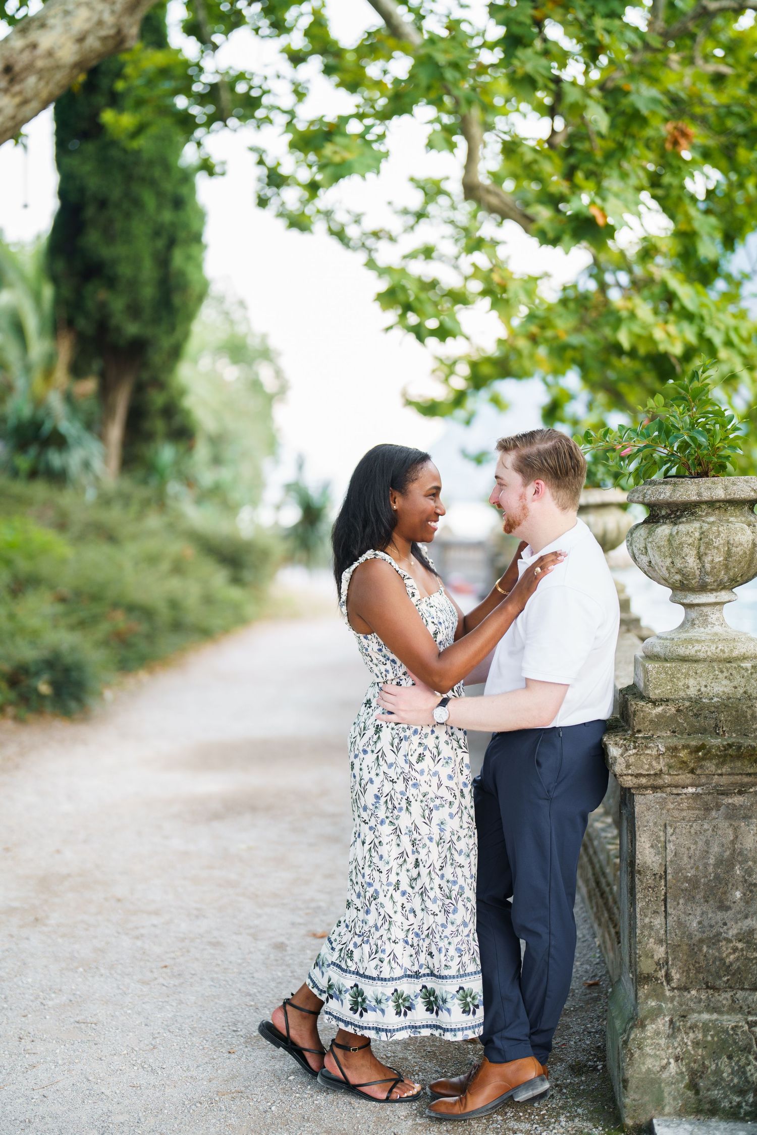 Loving interracial couple embracing and smiling by a stone urn on a lush garden pathway.