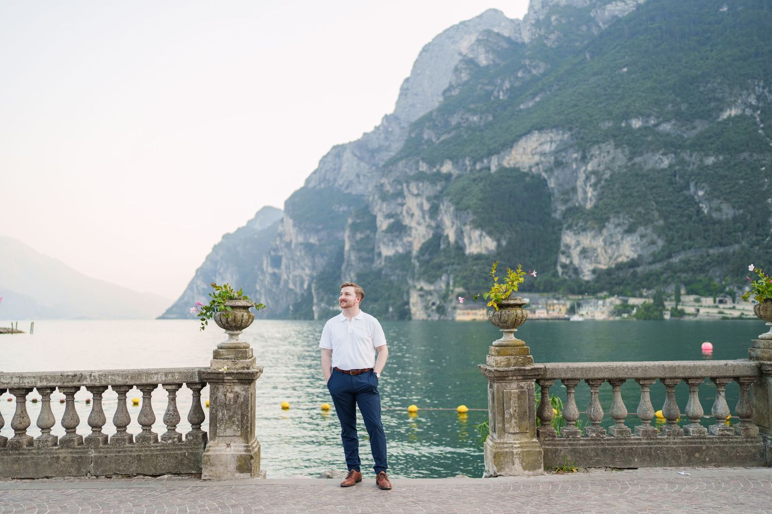 Well-dressed man posing on historic lakeside promenade with decorative urns and Lake Garda mountains behind.