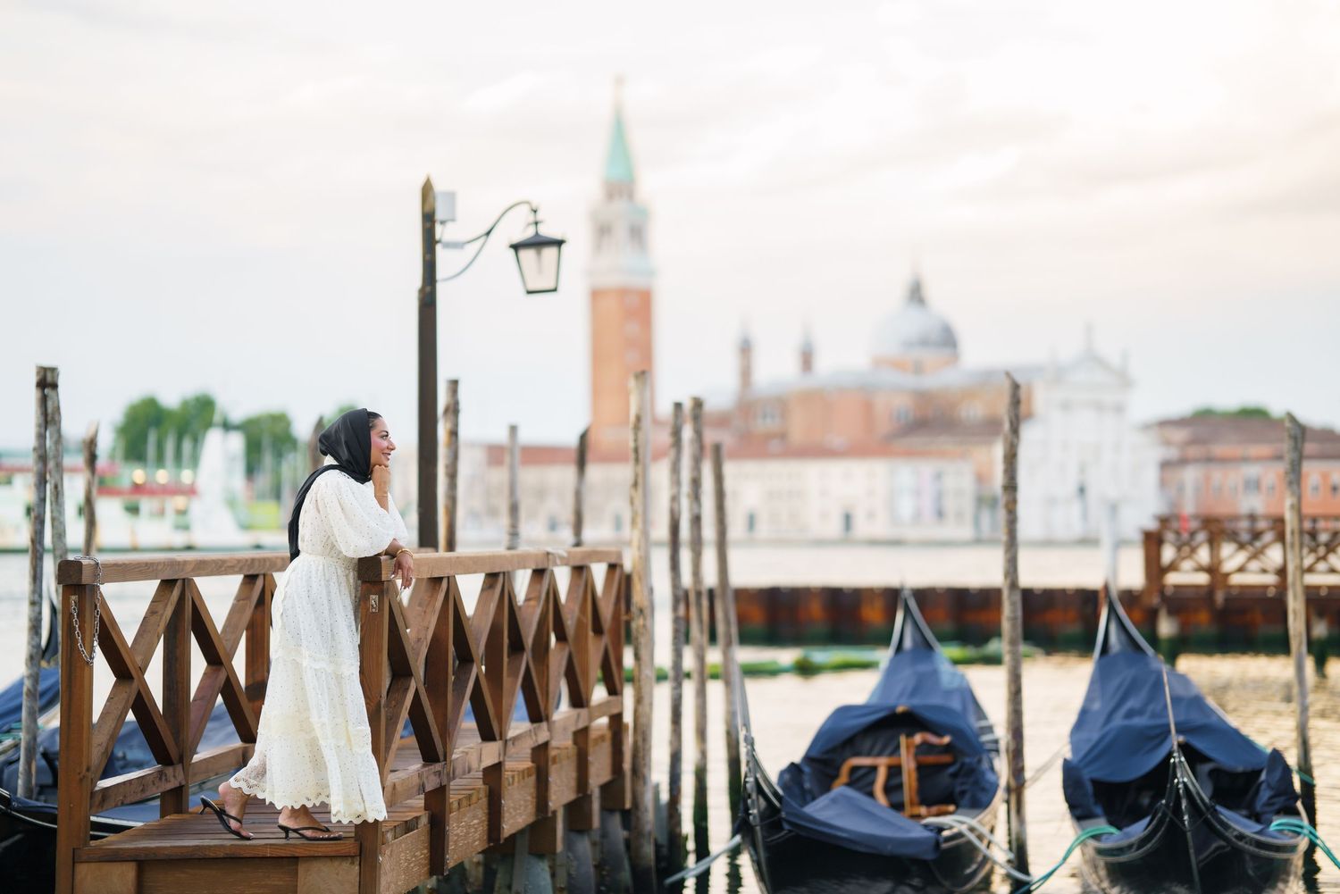 Woman in white dress standing on Venice dock with gondolas and San Giorgio Maggiore church in background.