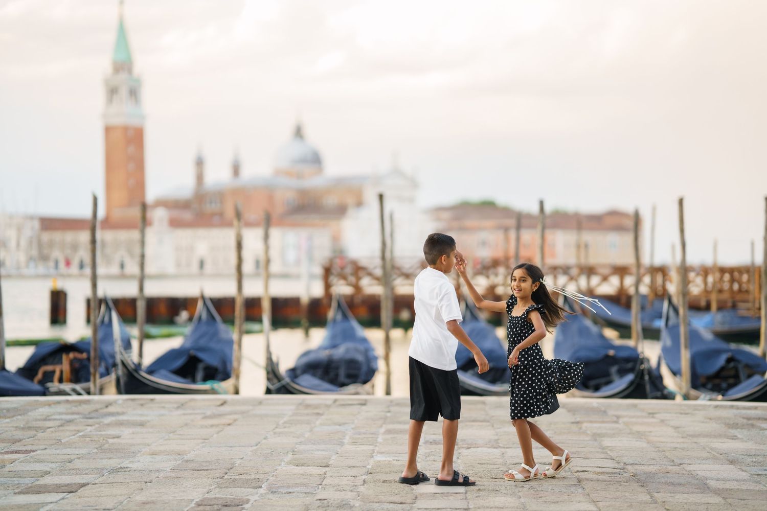 Couple holding hands by Venice gondolas with San Giorgio Maggiore church in background at sunset.