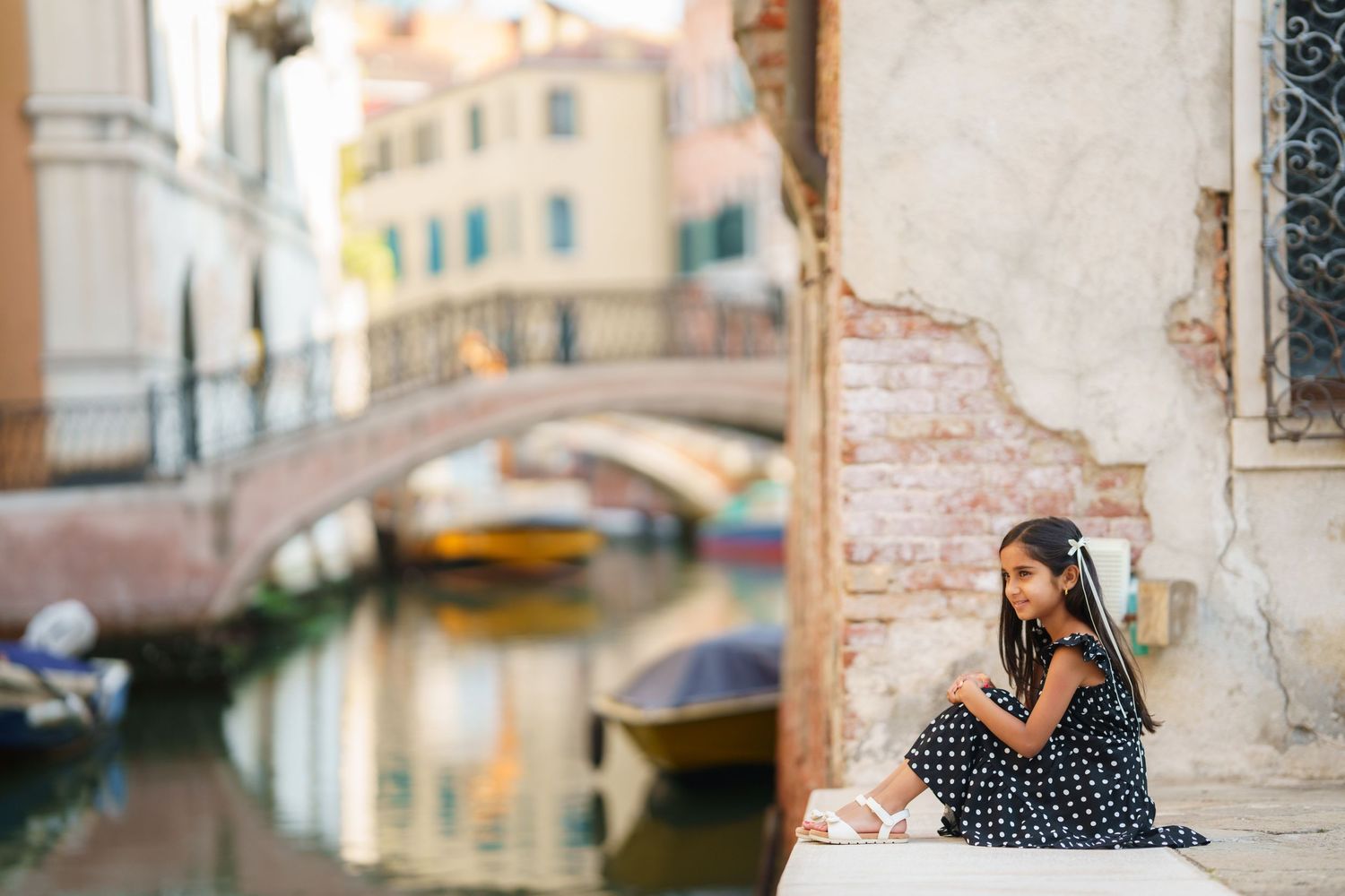Young girl in polka dot dress sitting by a Venice canal with bridge and colorful boats in background.