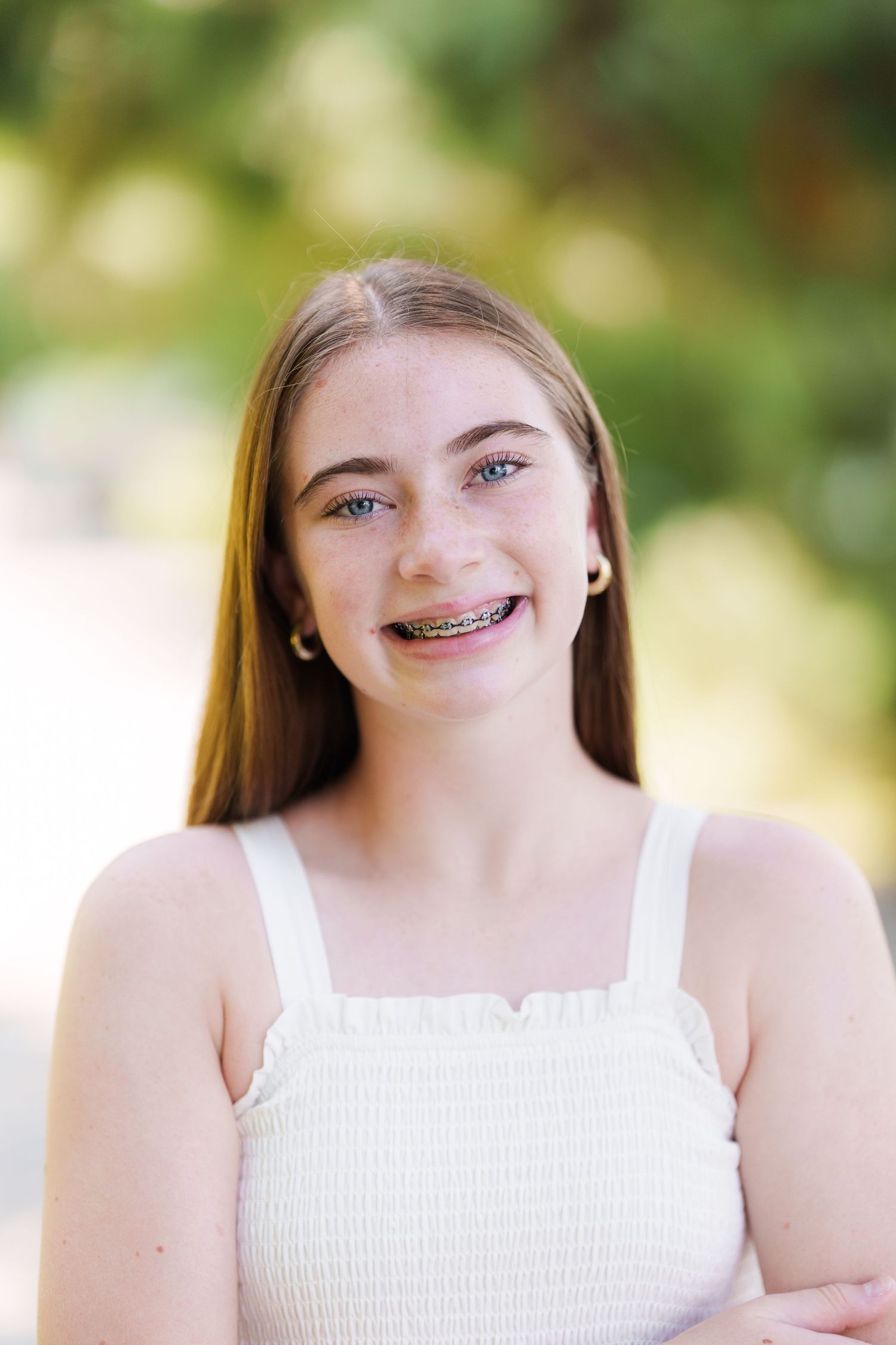 Smiling teenage girl with braces and long brown hair wearing a white top outdoors.