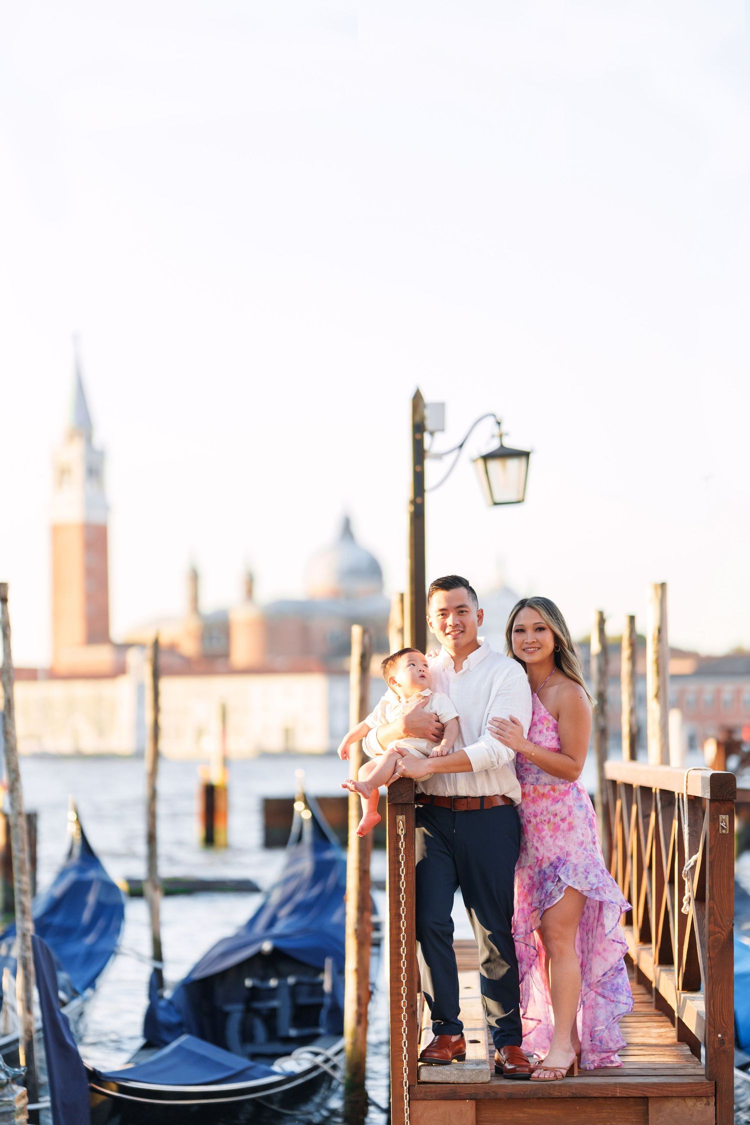 Family with baby posing on Venice dock with gondolas and San Giorgio Maggiore church in background.