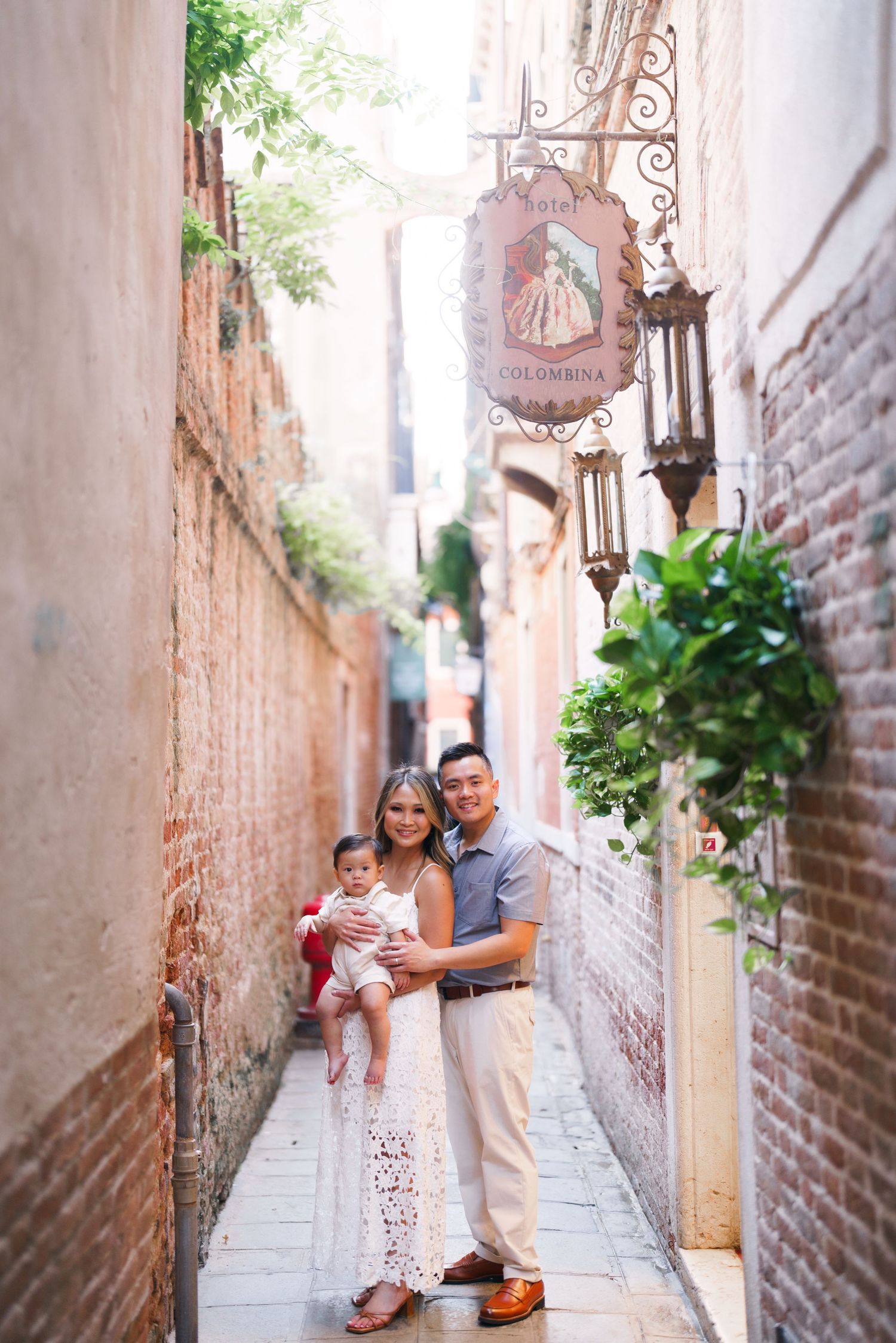 Family of three posing in a narrow Venice alley with brick walls, a decorative sign, and lush greenery.