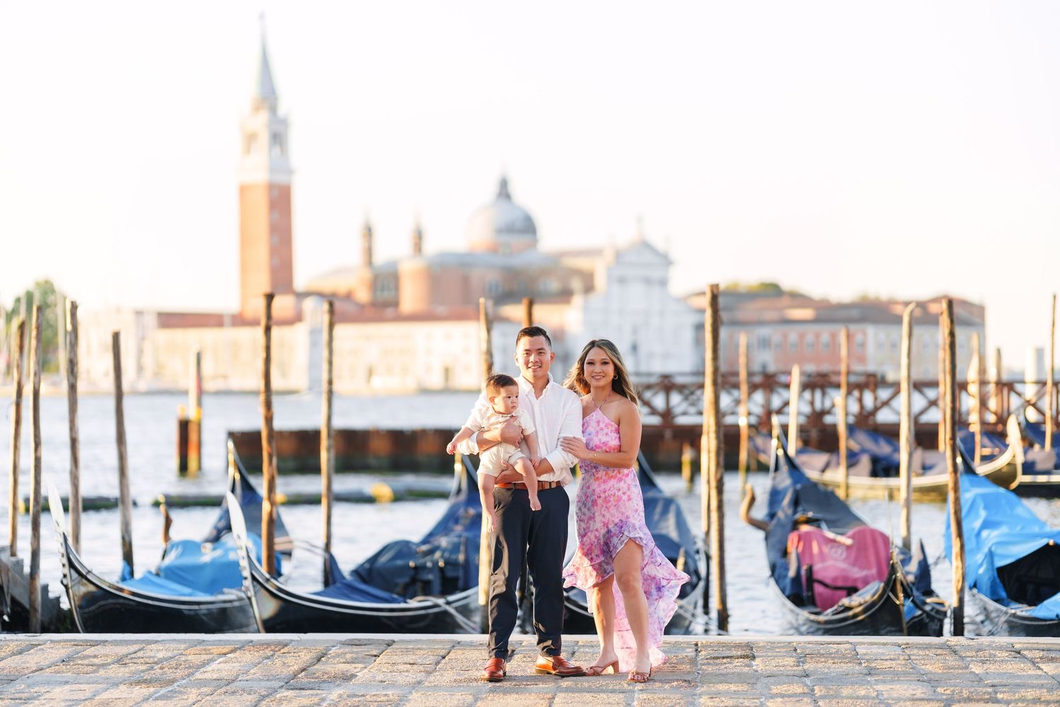 Family posing by gondolas in Venice, Italy, with San Giorgio Maggiore church visible across the lagoon.