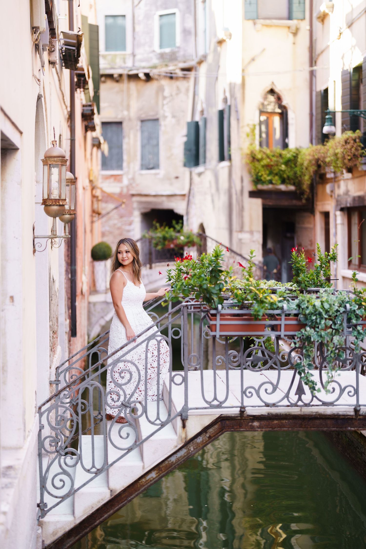 Bride in white dress standing on ornate iron bridge over Venice canal surrounded by colorful flowers and historic buildings.