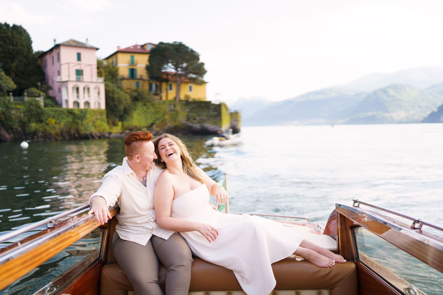 Couple laughing on a wooden boat on Lake Como, Italy, with colorful villas and mountains in the background.