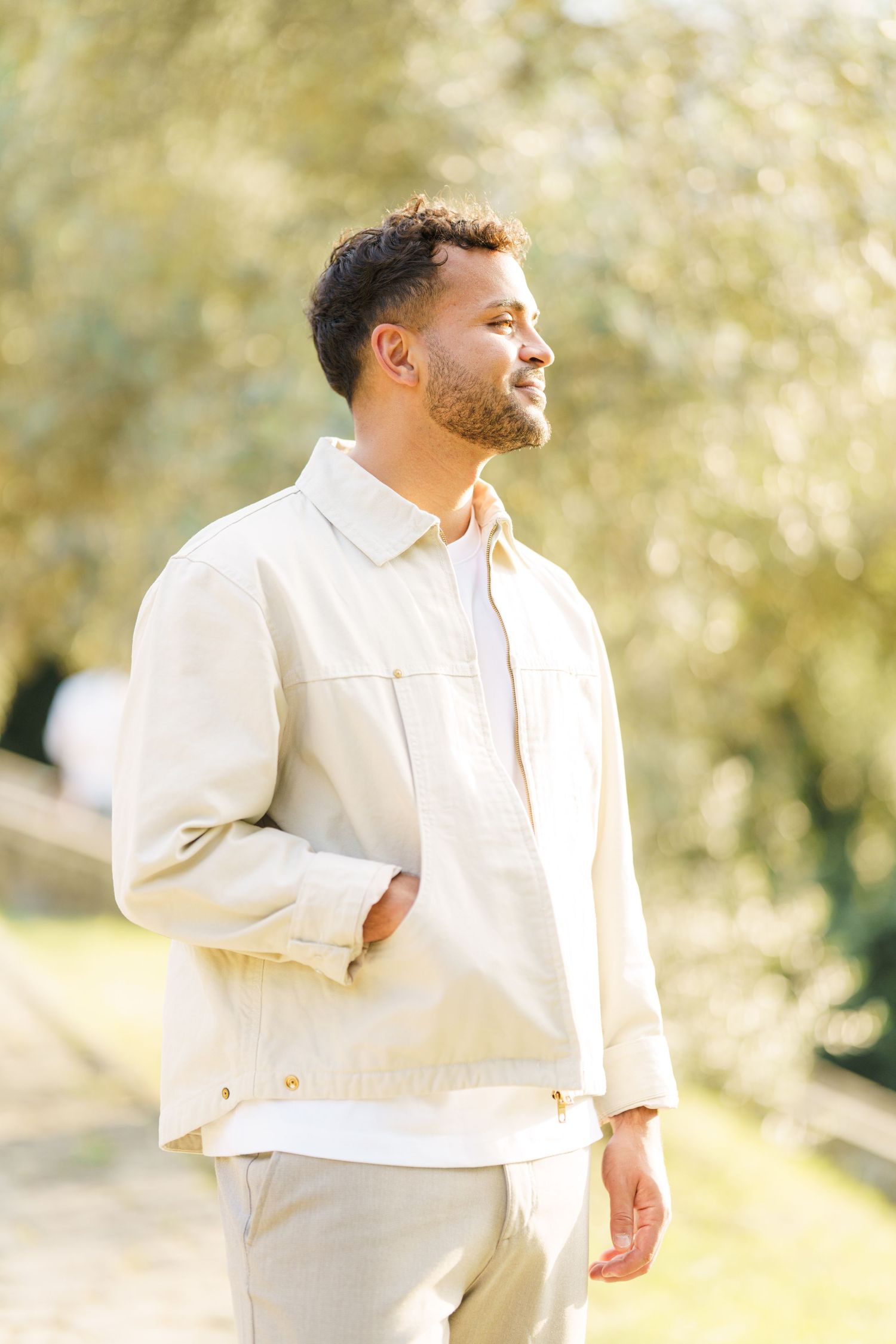 A young bearded man in a cream jacket and pants stands outdoors in a sunny park, gazing thoughtfully to the side.