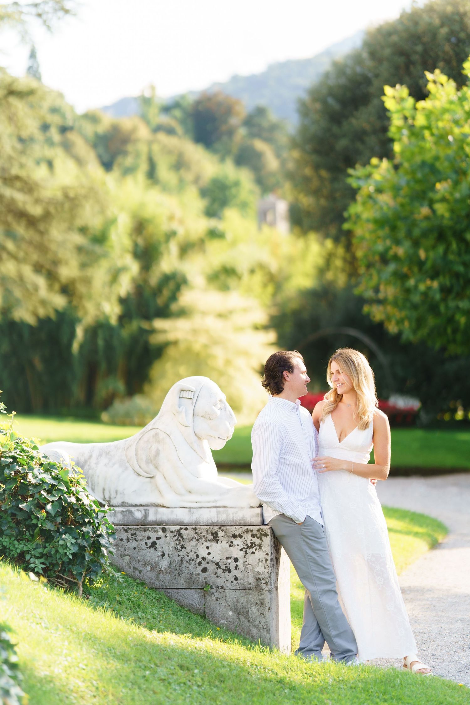 A romantic couple in white attire share intimate moments on a stone wall in a sunlit garden setting.