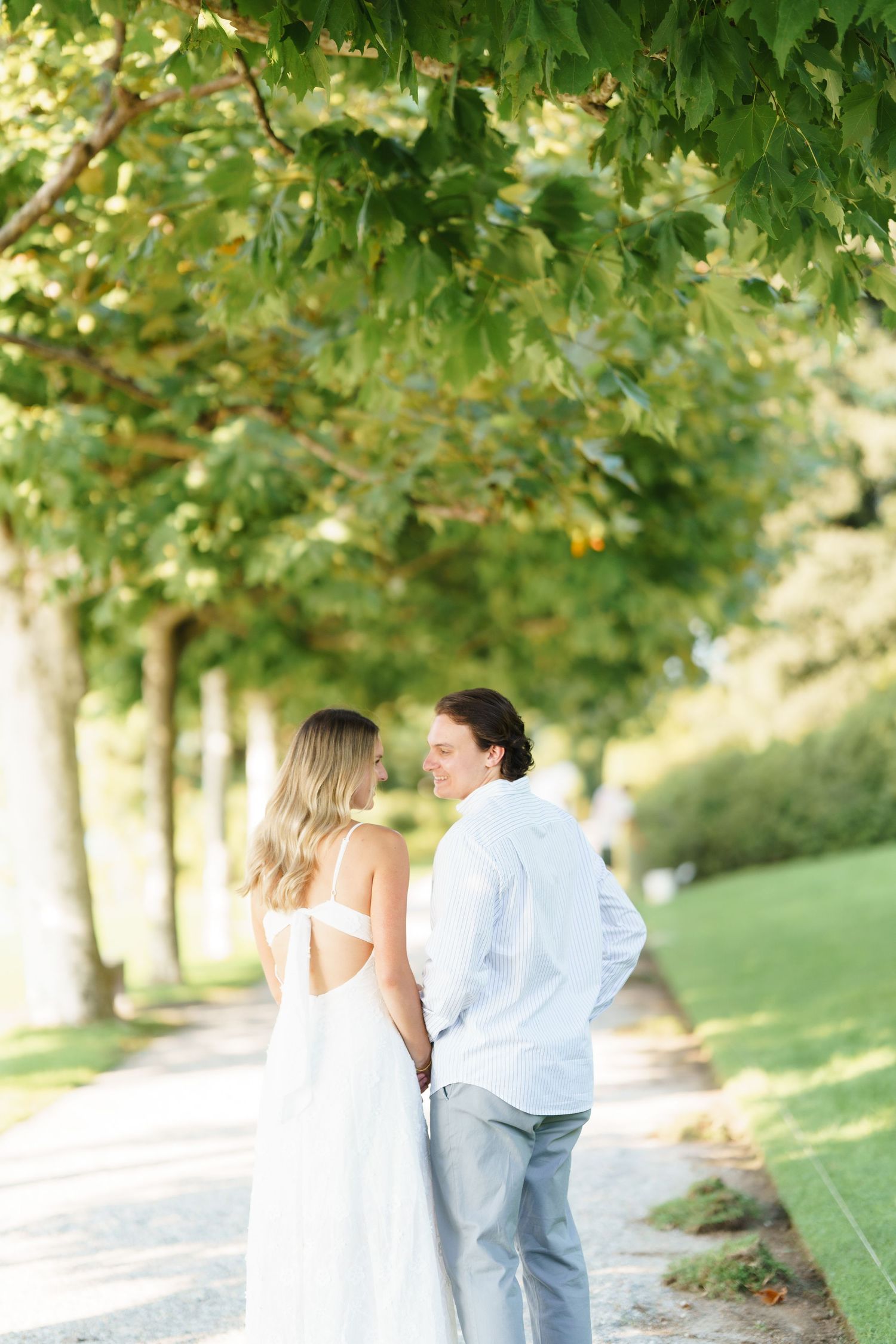 A couple in white and light blue attire walks along a tree-lined path on a sunny summer day.
